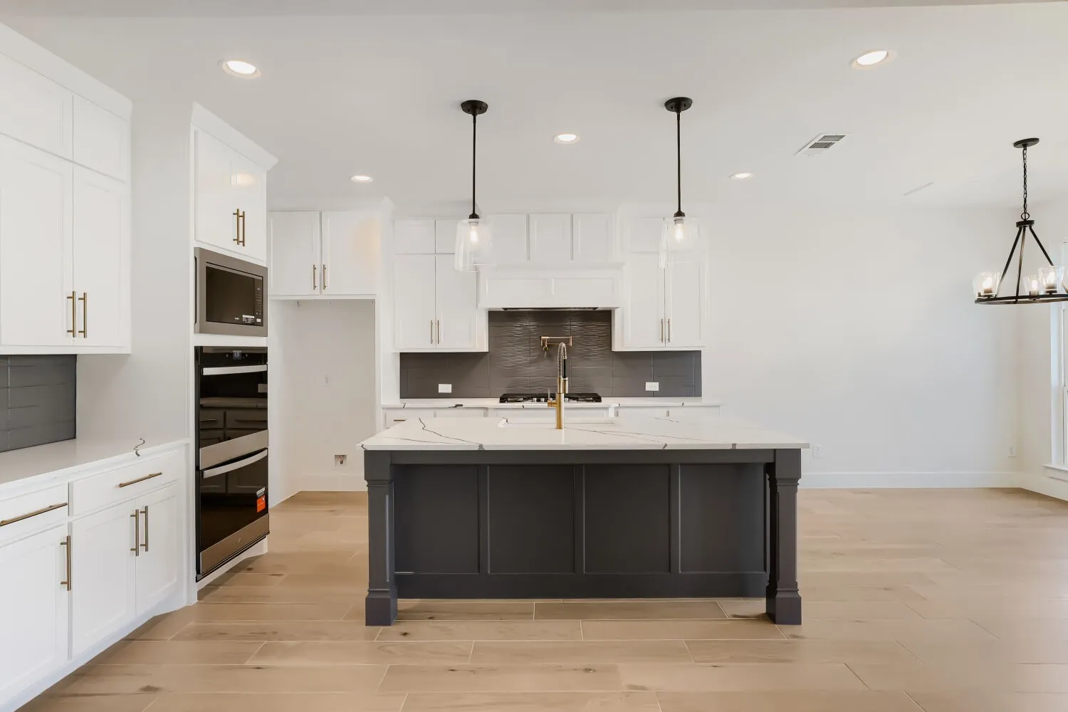 Kitchen featuring a dark-toned island with a light-toned countertop, white cabinetry with gold-toned hardware, built-in stainless steel oven and microwave, light wood-finish flooring, and recessed lighting