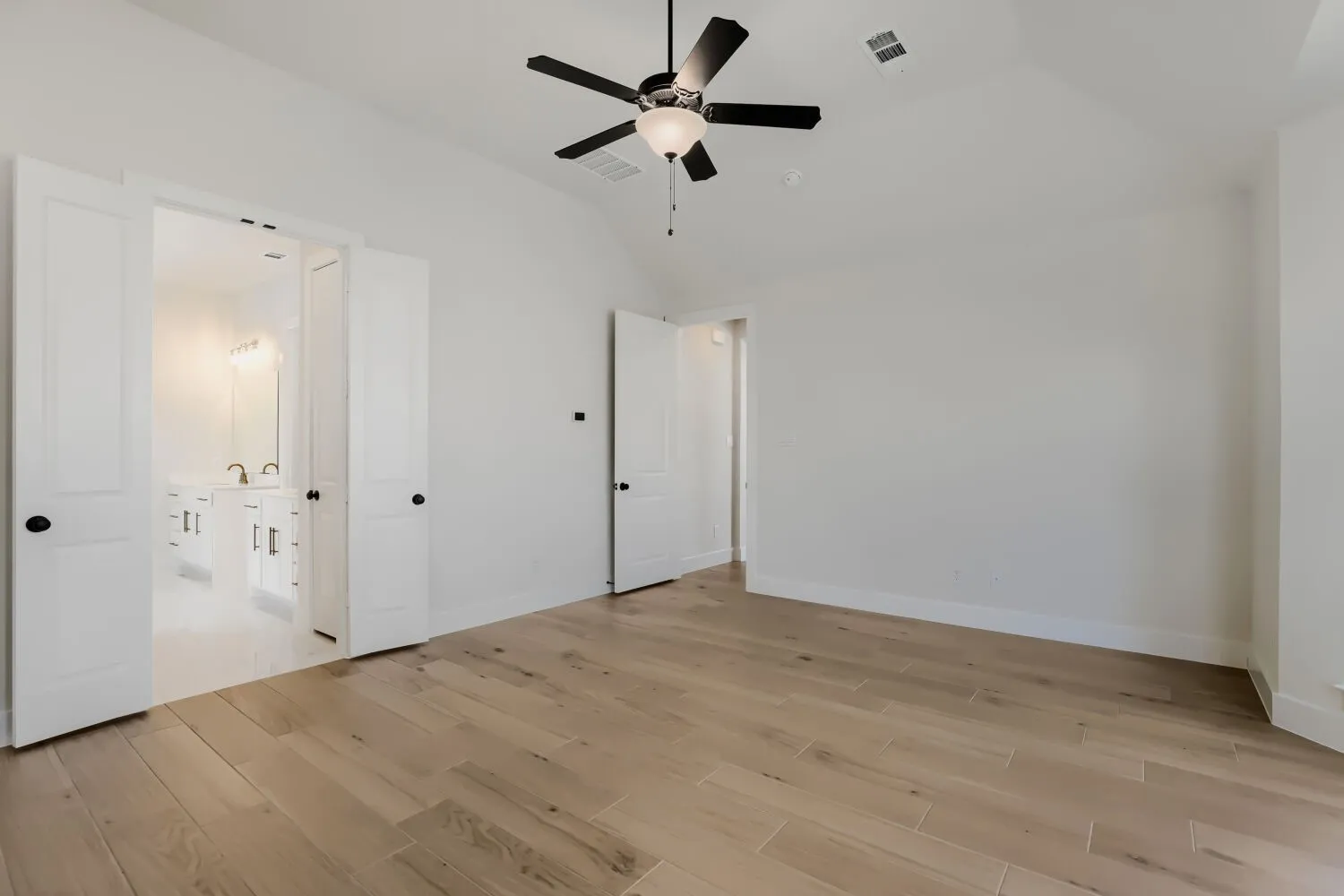 Vaulted ceiling with a black ceiling fan, wood-finish flooring, and white double doors opening to a bathroom with a dual vanity