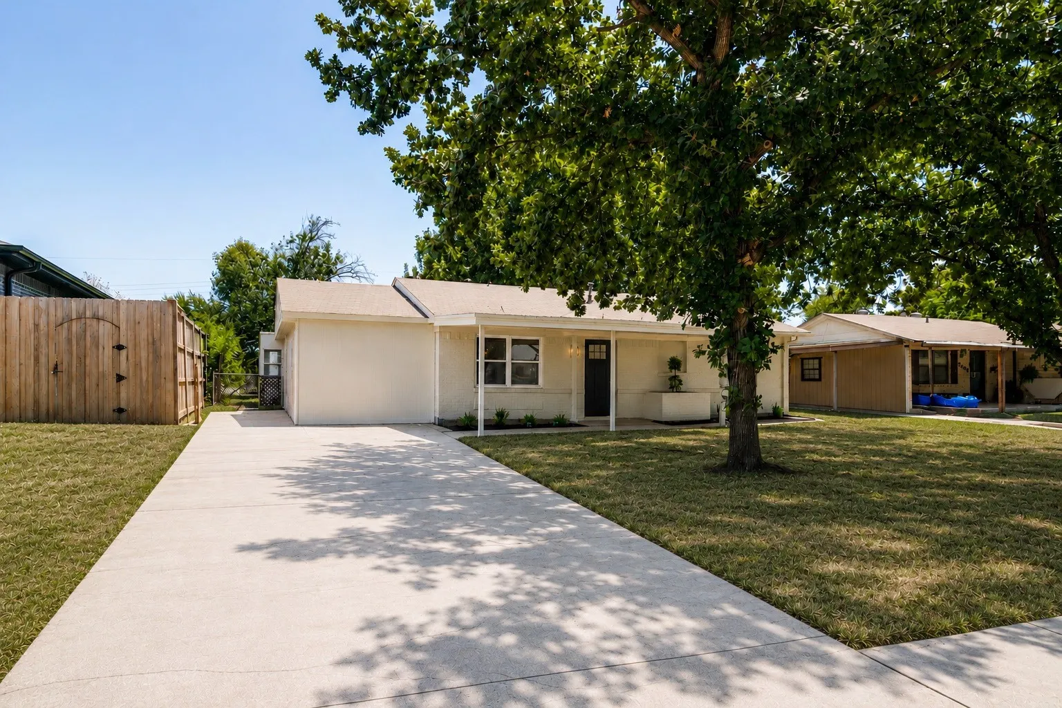 Spacious concrete driveway, manicured lawn, wood privacy fence, covered front porch, and mature shade trees