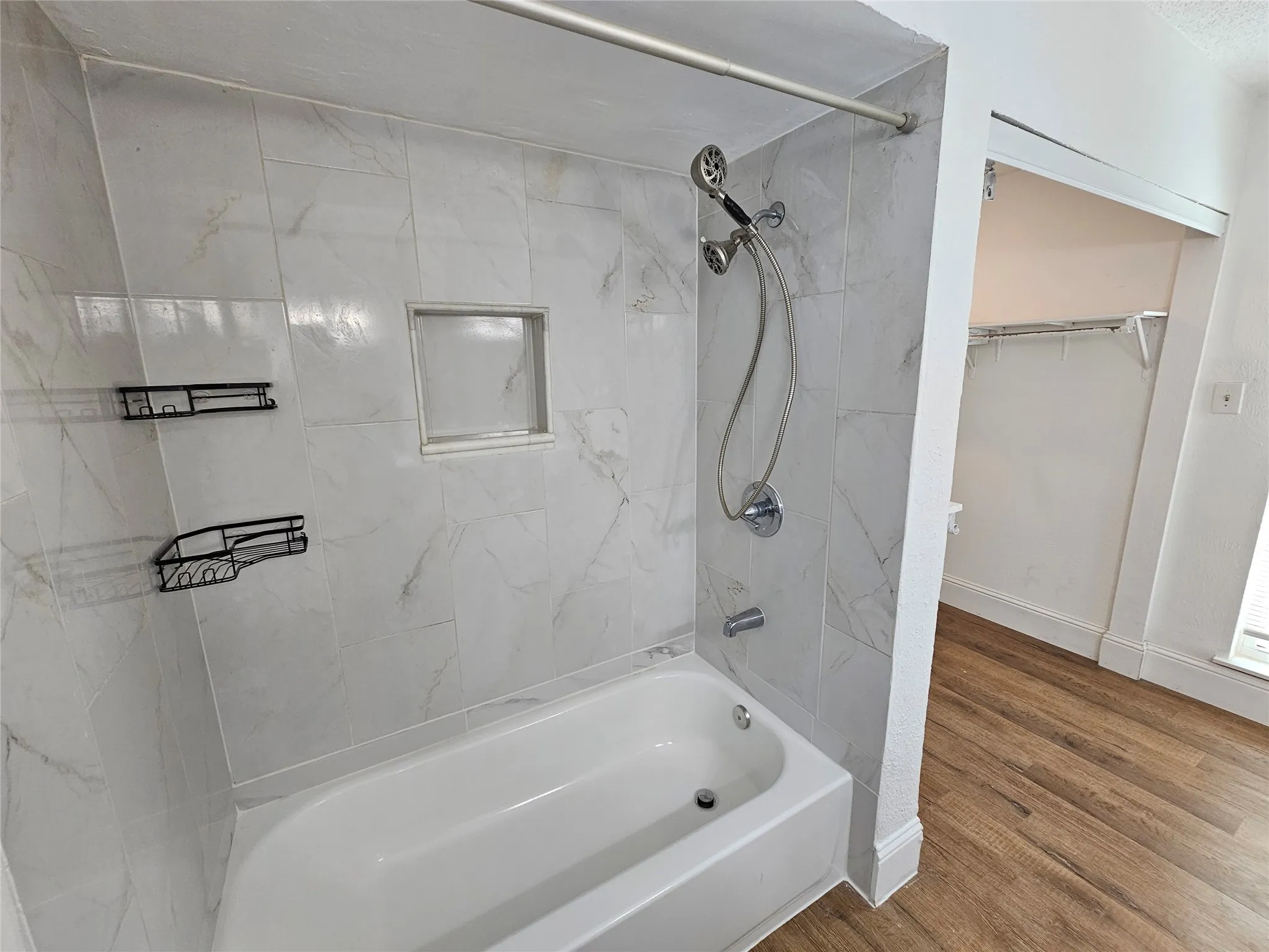 Bathtub with tiled surround, recessed shower niche, wall-mounted shower fixture, and wood-finish flooring