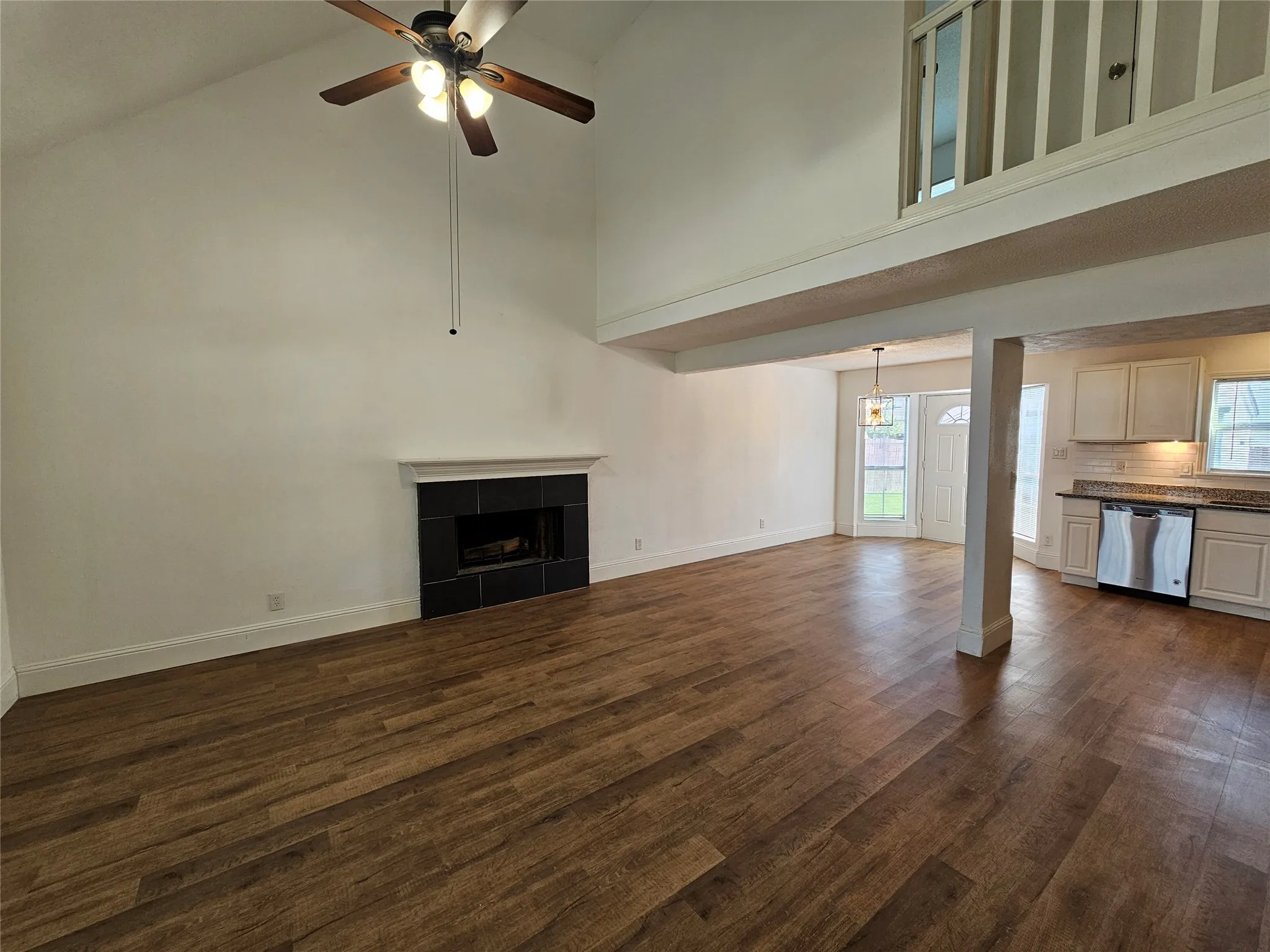 Vaulted ceiling with a ceiling fan, fireplace with a black tile surround, wood-finish flooring, and a kitchen with light-colored cabinetry and stainless steel appliances
