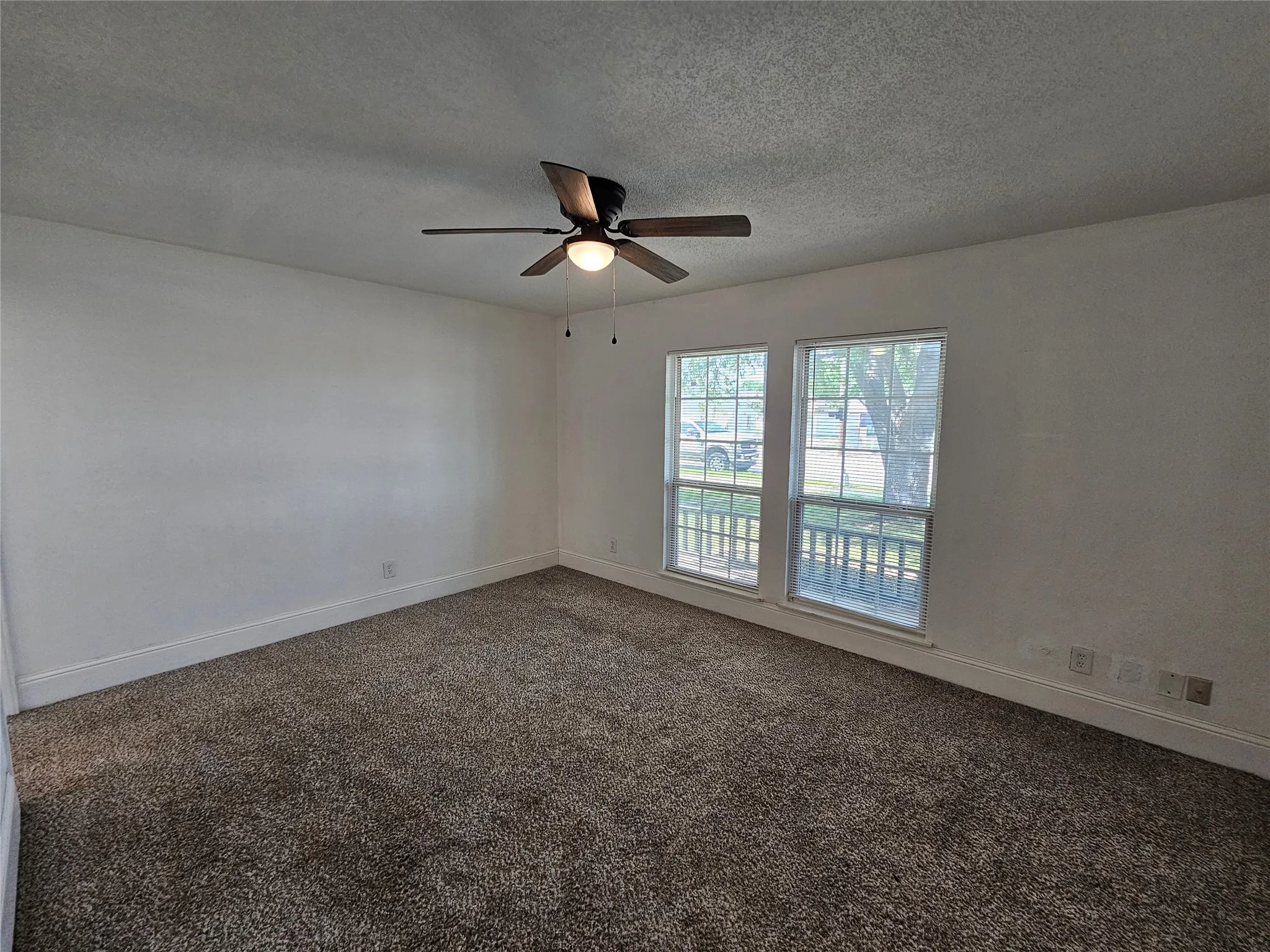 Neutral wall color with textured carpeting, featuring two large windows and a ceiling fan with integrated lighting