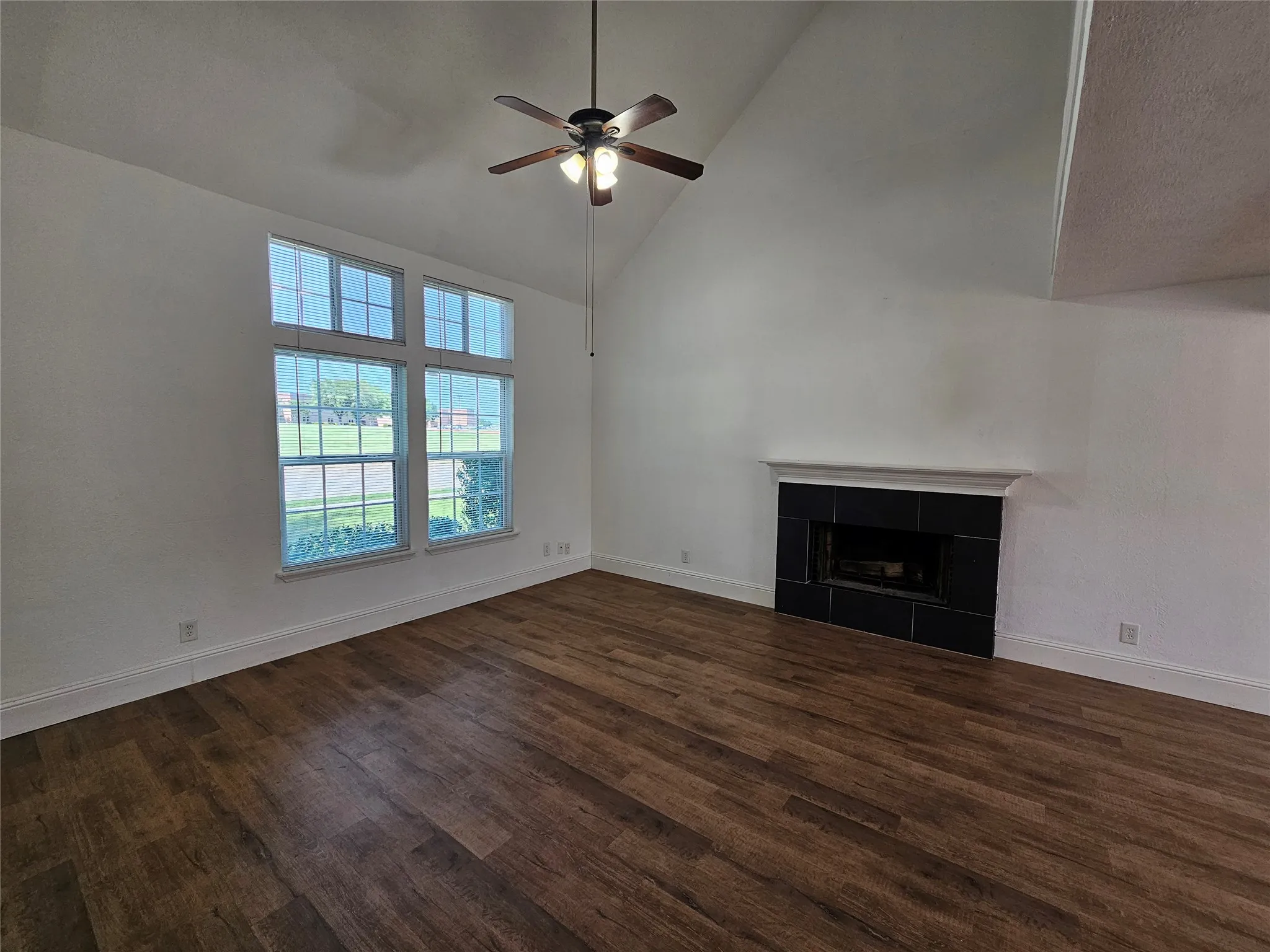 Vaulted ceilings, wood-finish flooring, a ceiling fan with integrated lighting, a fireplace with a tiled surround, and large windows