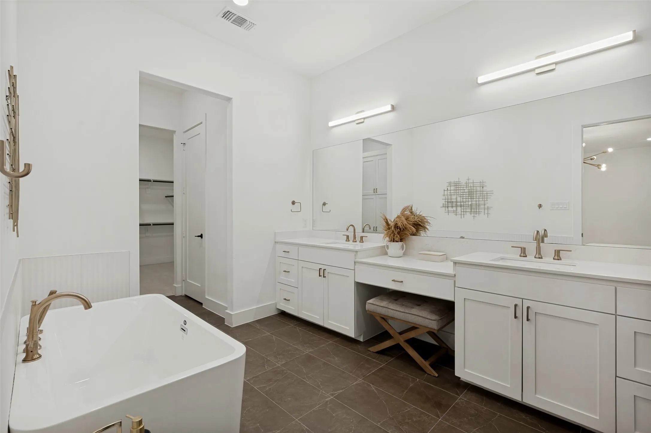 Freestanding soaking tub with gold-tone fixtures, dual white vanity cabinets with integrated sinks, expansive wall-mounted mirror, and tile flooring