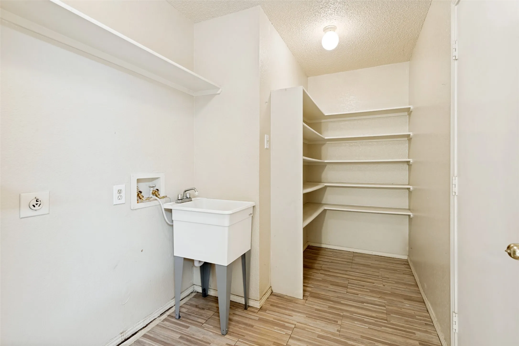 Utility room with a laundry basin, overhead shelving, and wood-finish flooring