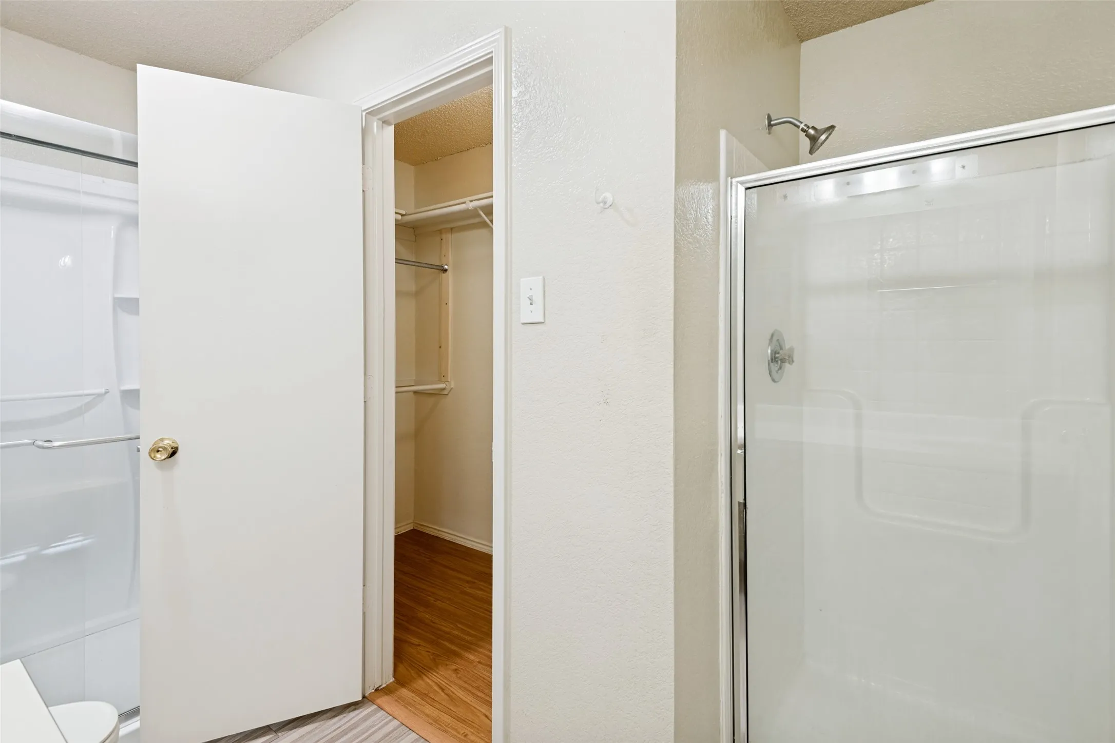 White interior door with brass hardware, walk-in closet with wood-finish flooring and shelving, and a shower with a sliding glass door and chrome fixtures