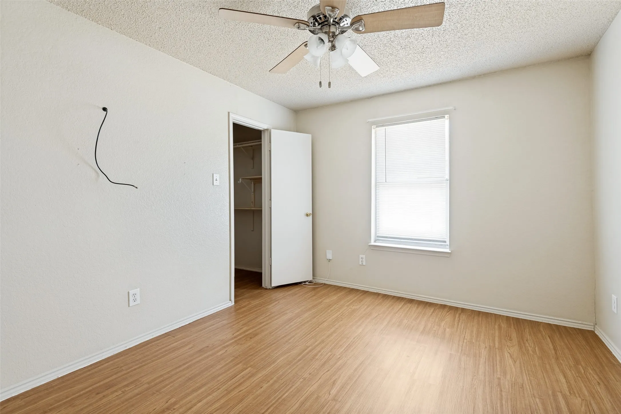 Wood-finish flooring with white baseboards, window with blinds, ceiling fan, and an open closet with shelving