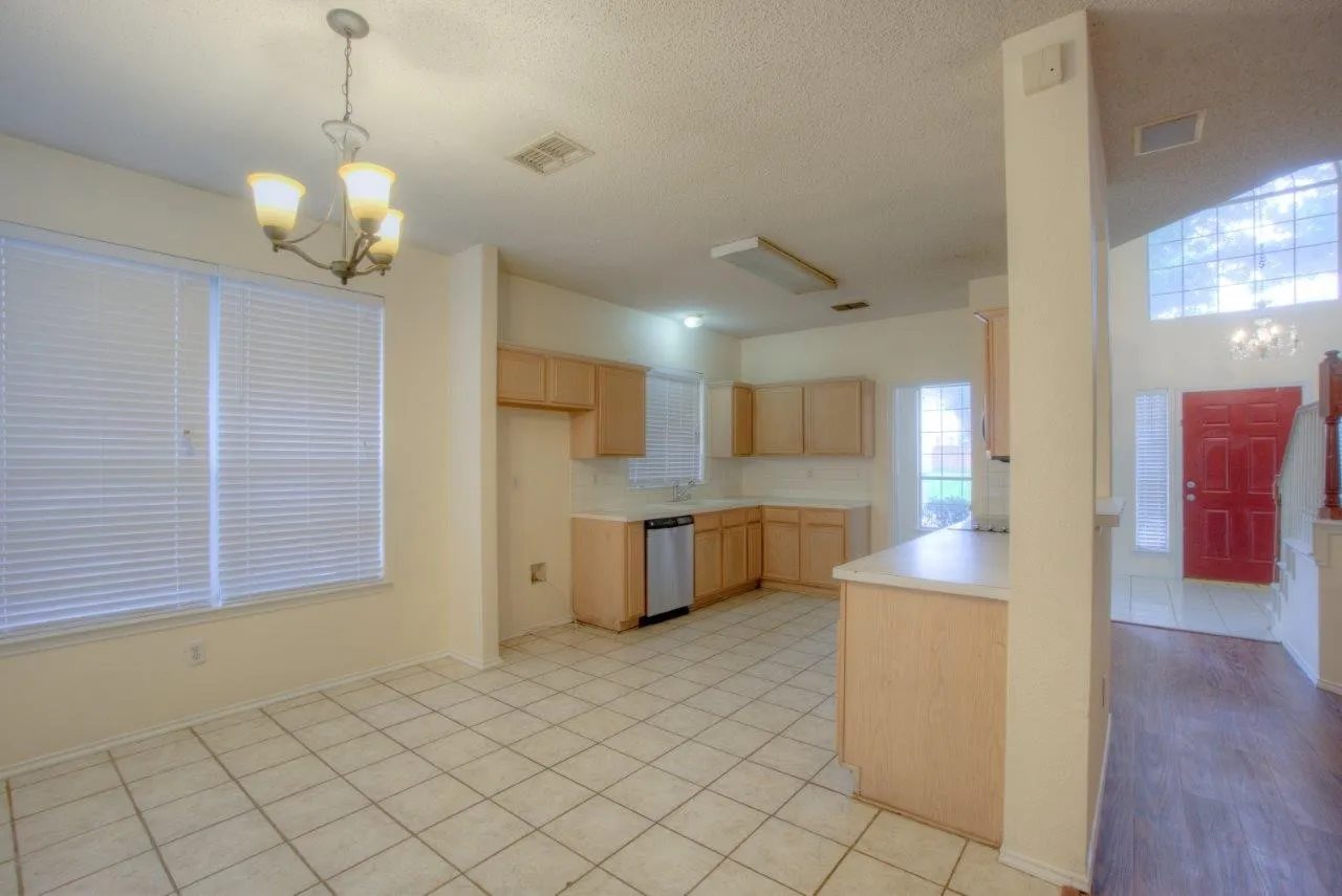 Kitchen and dining area featuring tile flooring, light wood-finish cabinetry, stainless steel dishwasher, a window with blinds, and a chandelier light fixture