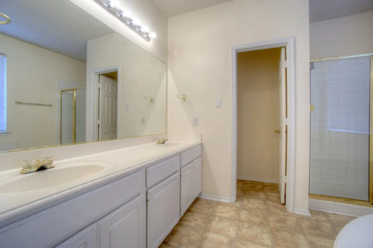 Double vanity with integrated sinks, full-width mirror, overhead bar lighting, and a shower enclosure with a gold-tone frame