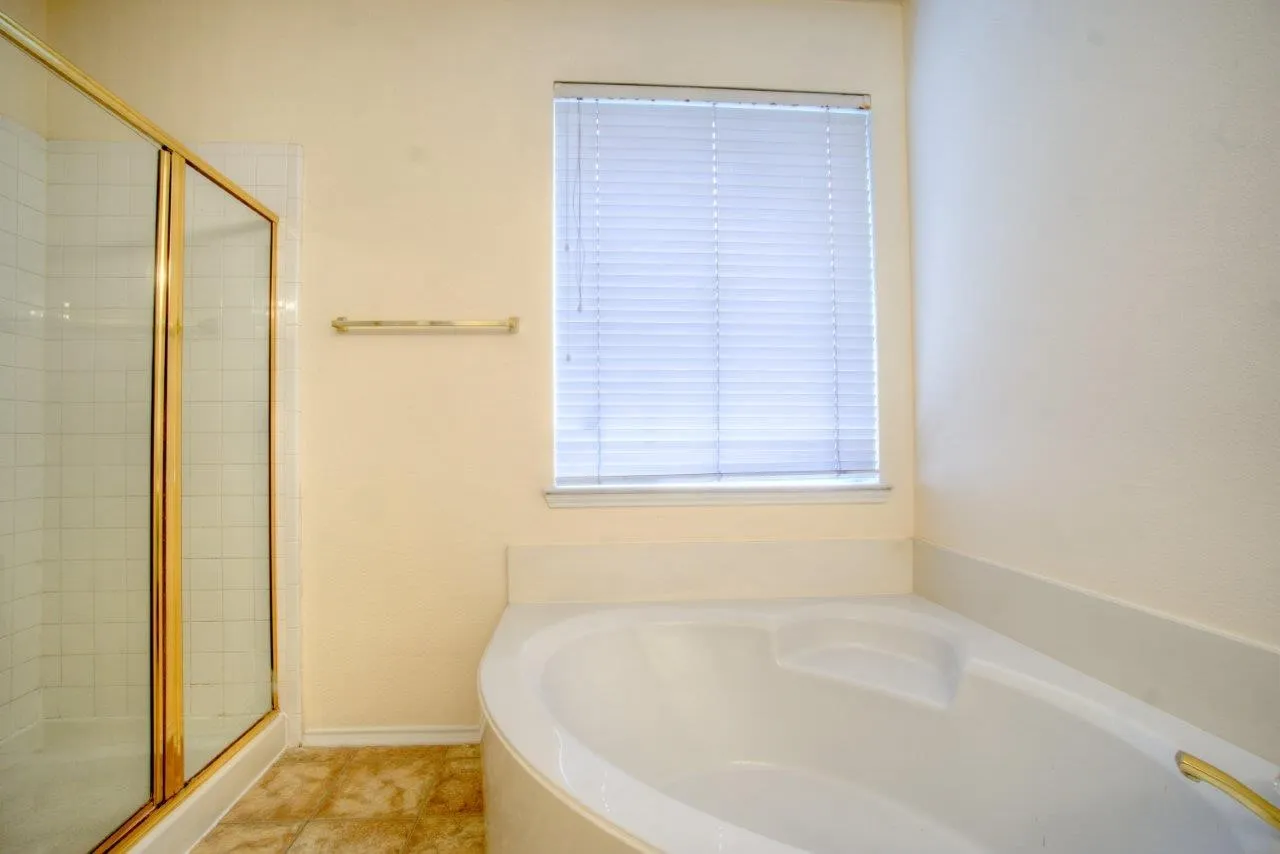 White corner soaking tub with integrated seating, adjacent to a shower enclosure with a golden frame and white tile surround