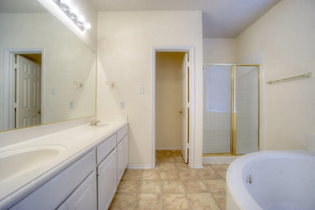 Dual sink vanity with white cabinetry, gold-framed shower enclosure, built-in bathtub, and tiled flooring