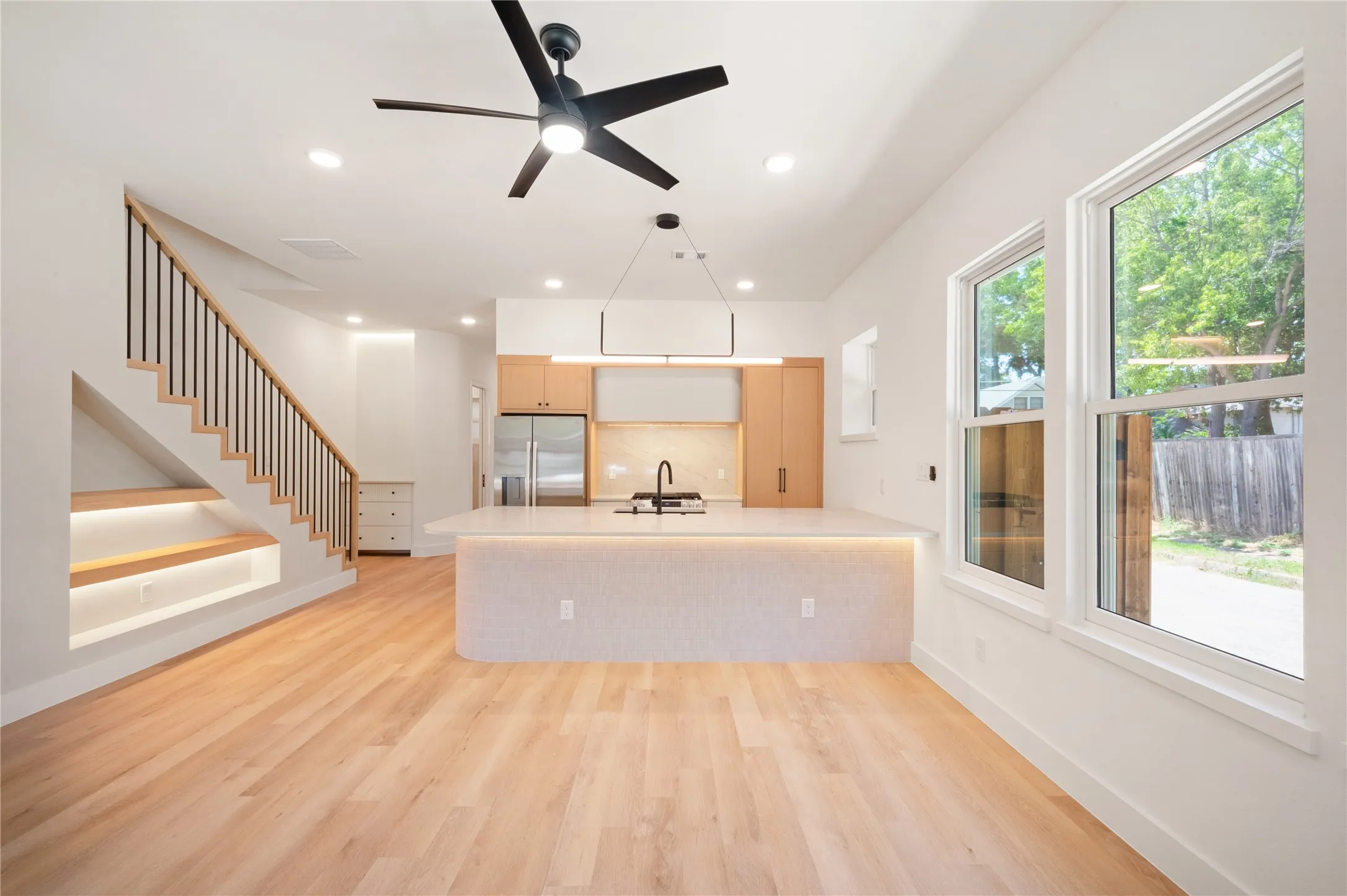 Light wood-finish flooring, staircase with black metal balusters, integrated under-stair shelving with lighting, kitchen island with integrated lighting and a white countertop, and expansive windows