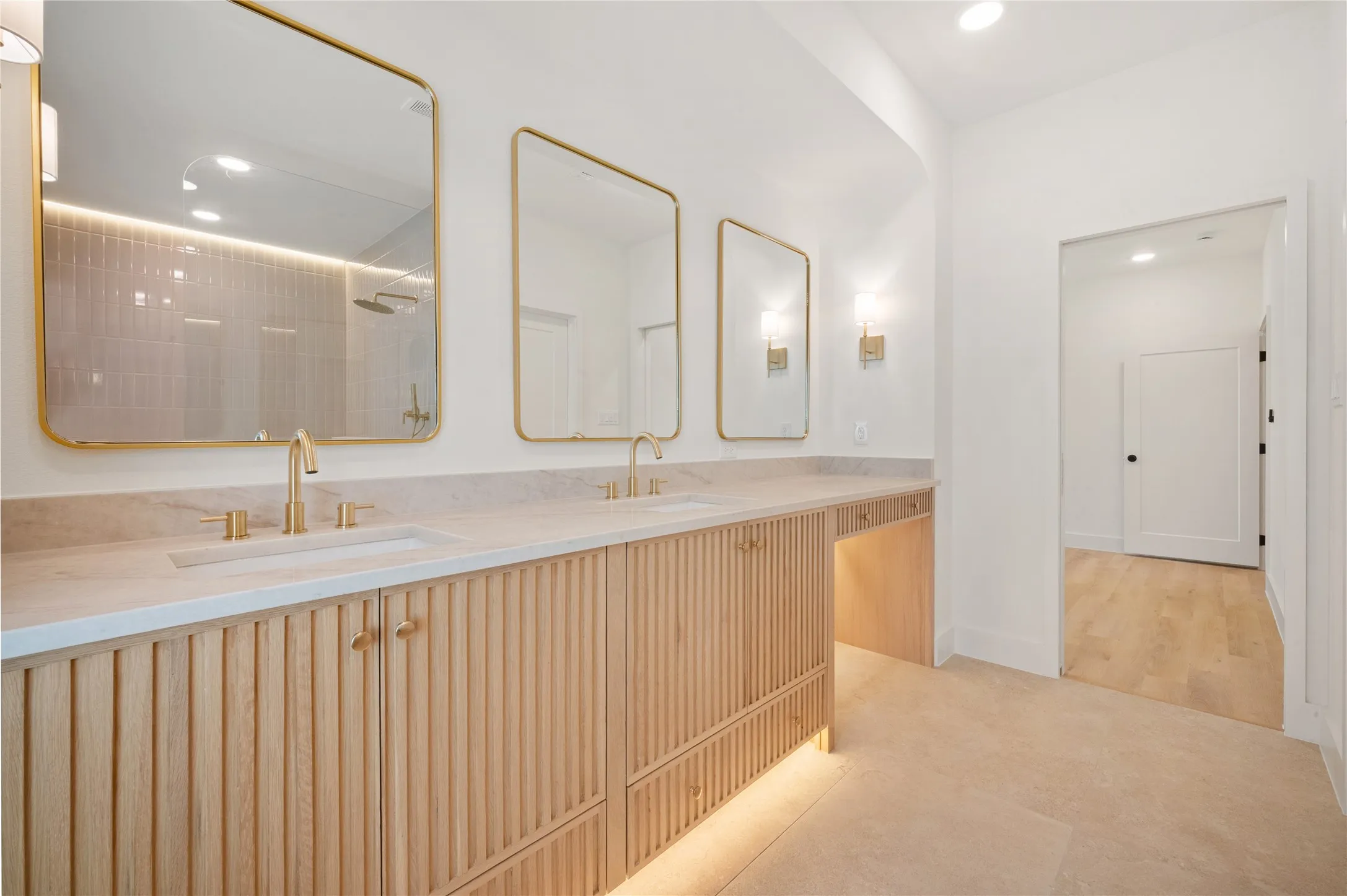 Dual vanity with integrated sinks, light wood-finish cabinetry with vertical fluting, light-toned countertop, and gold-framed mirrors