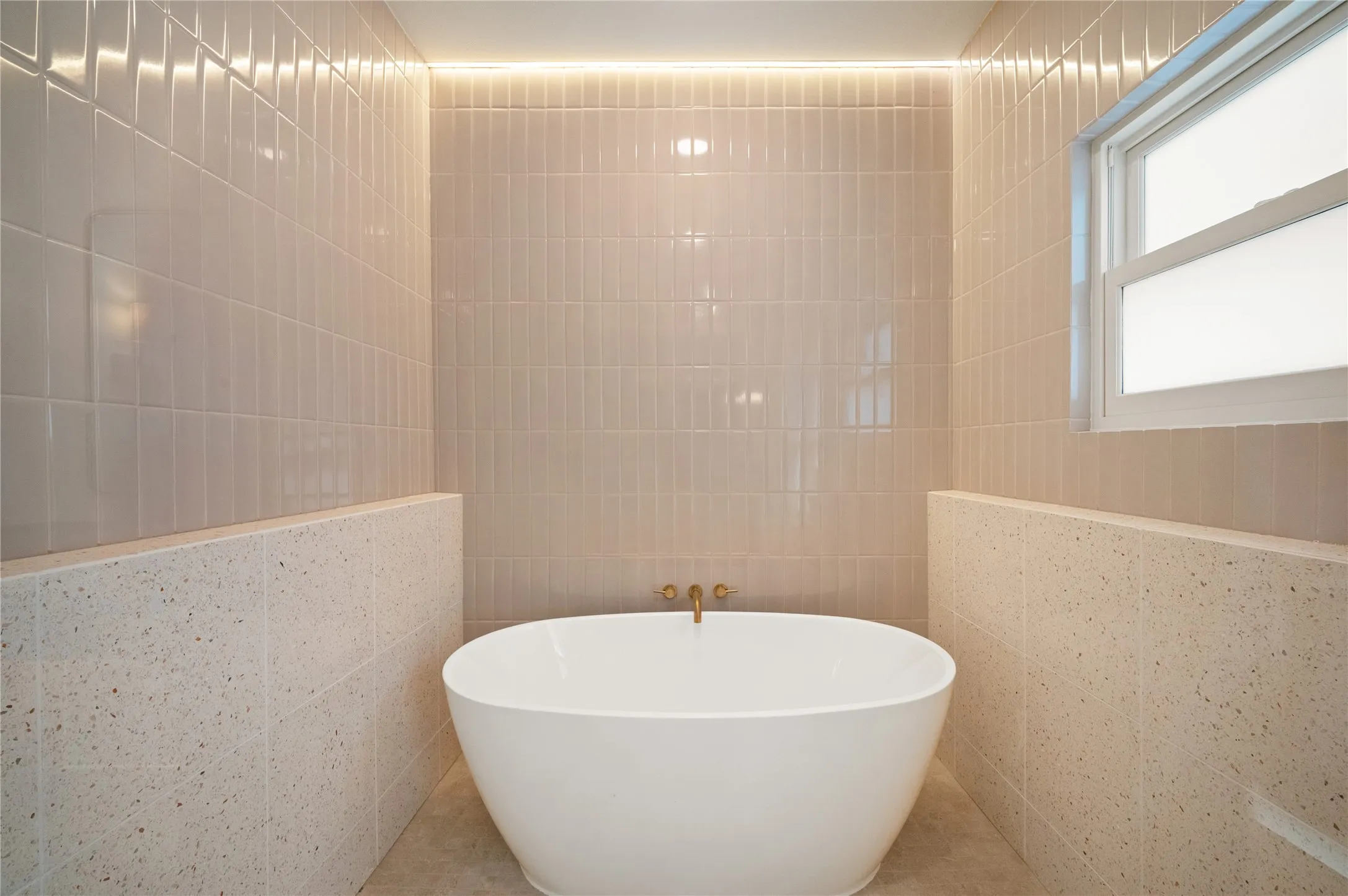 Freestanding soaking tub with a wall-mounted brass faucet, surrounded by tiled walls featuring a lower speckled pattern and upper vertical rectangular tiles