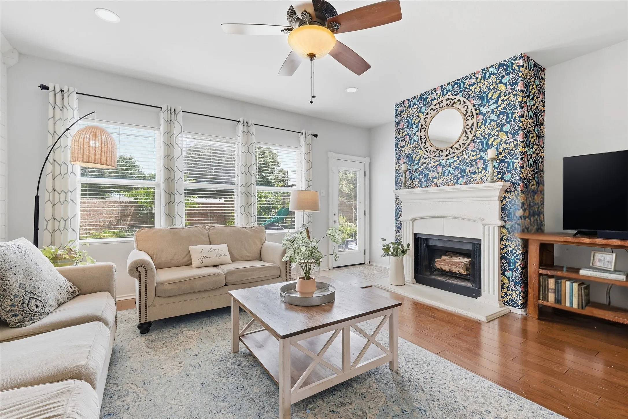 Living area featuring a floor-to-ceiling accent wall, an ornate fireplace with a white mantel, wood-finish flooring, a ceiling fan with light fixture, and multiple windows with blinds