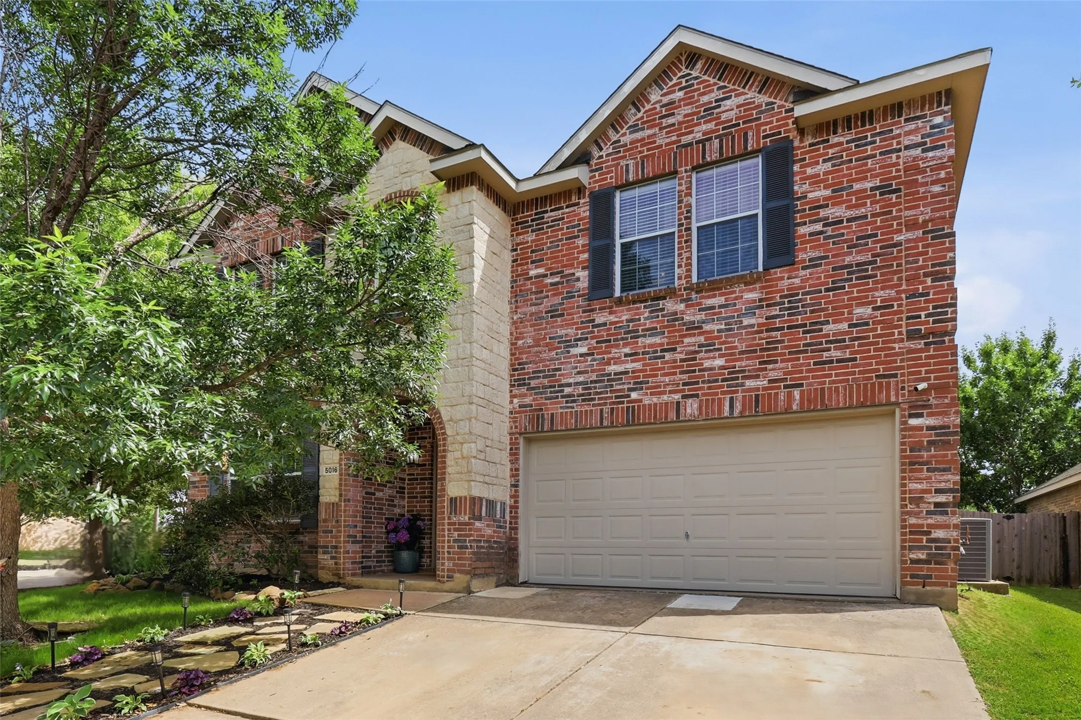 Brick and stone facade featuring a two-car garage, arched entryway, and mature landscaping