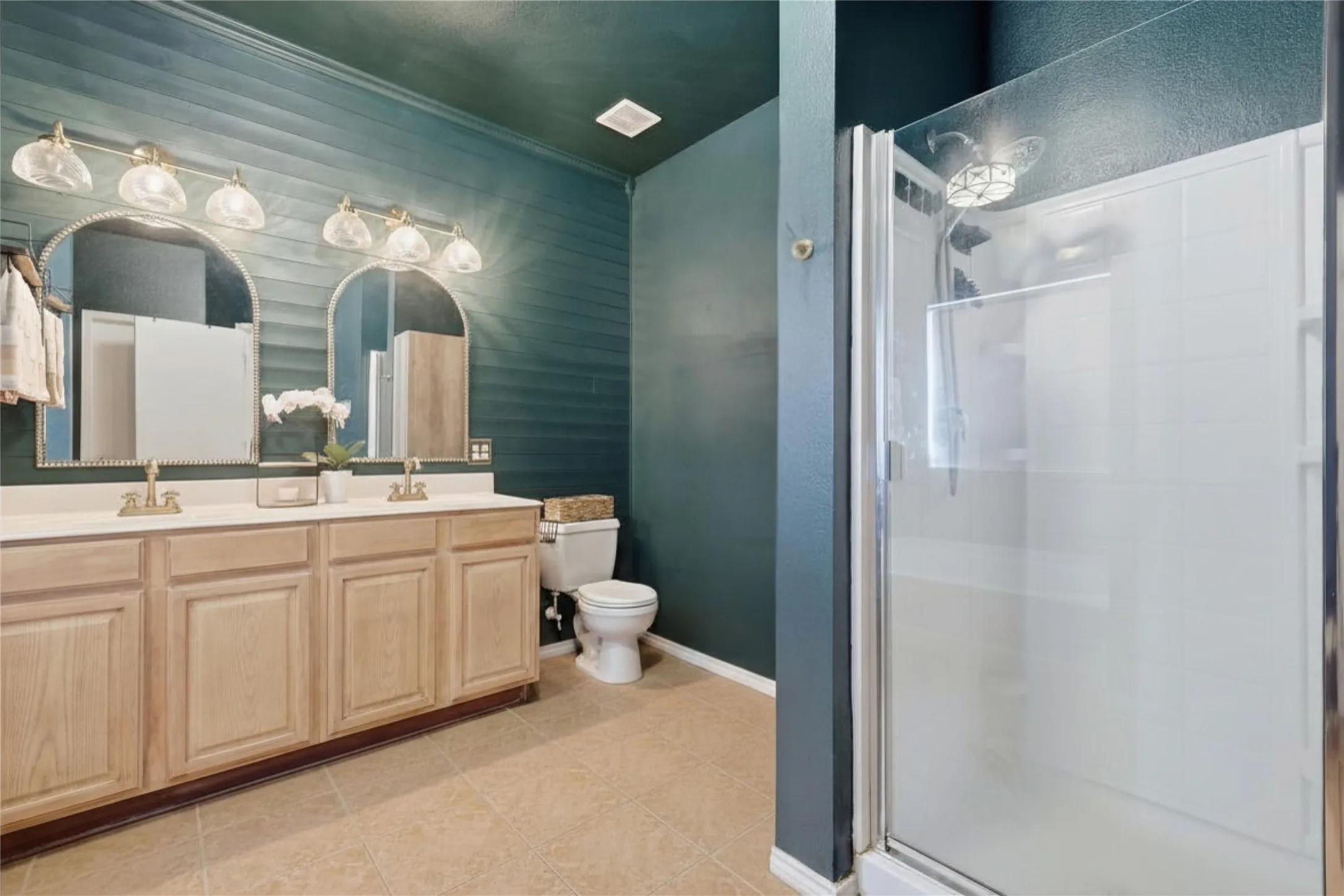 Bathroom featuring a dual vanity with light wood cabinetry, two arched mirrors, and a shower enclosure with a clear glass door