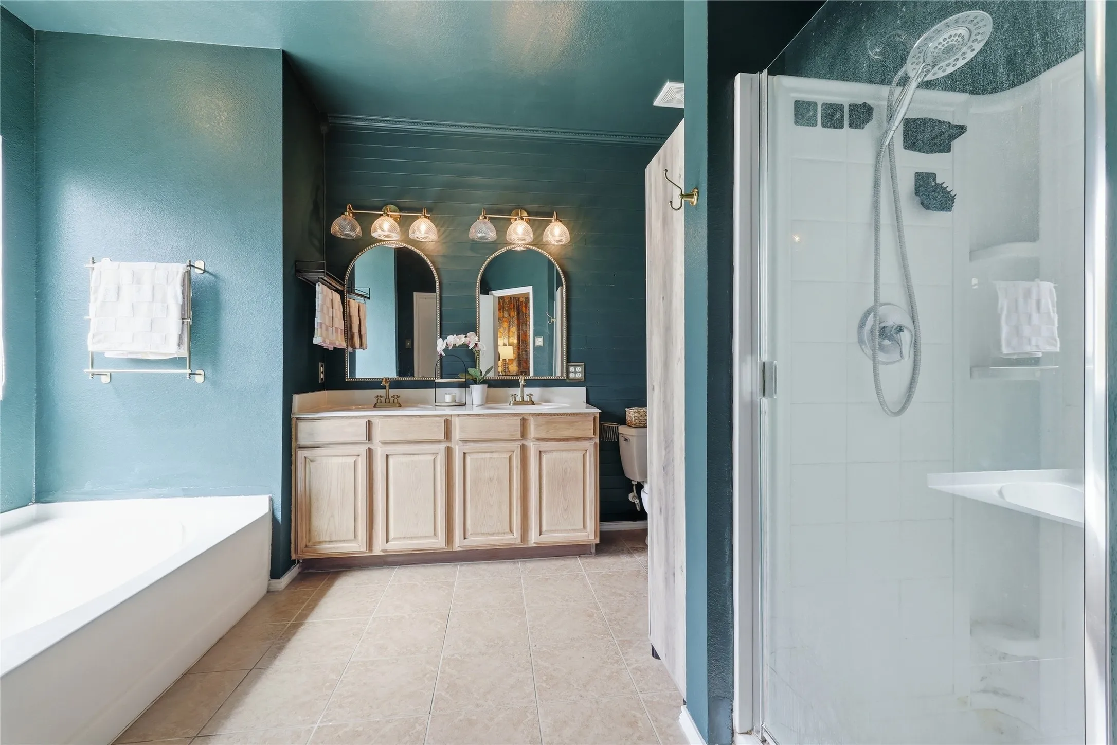 Dual vanity with arched mirrors, a built-in shower stall with glass enclosure, a white soaking tub, and neutral-toned tile flooring
