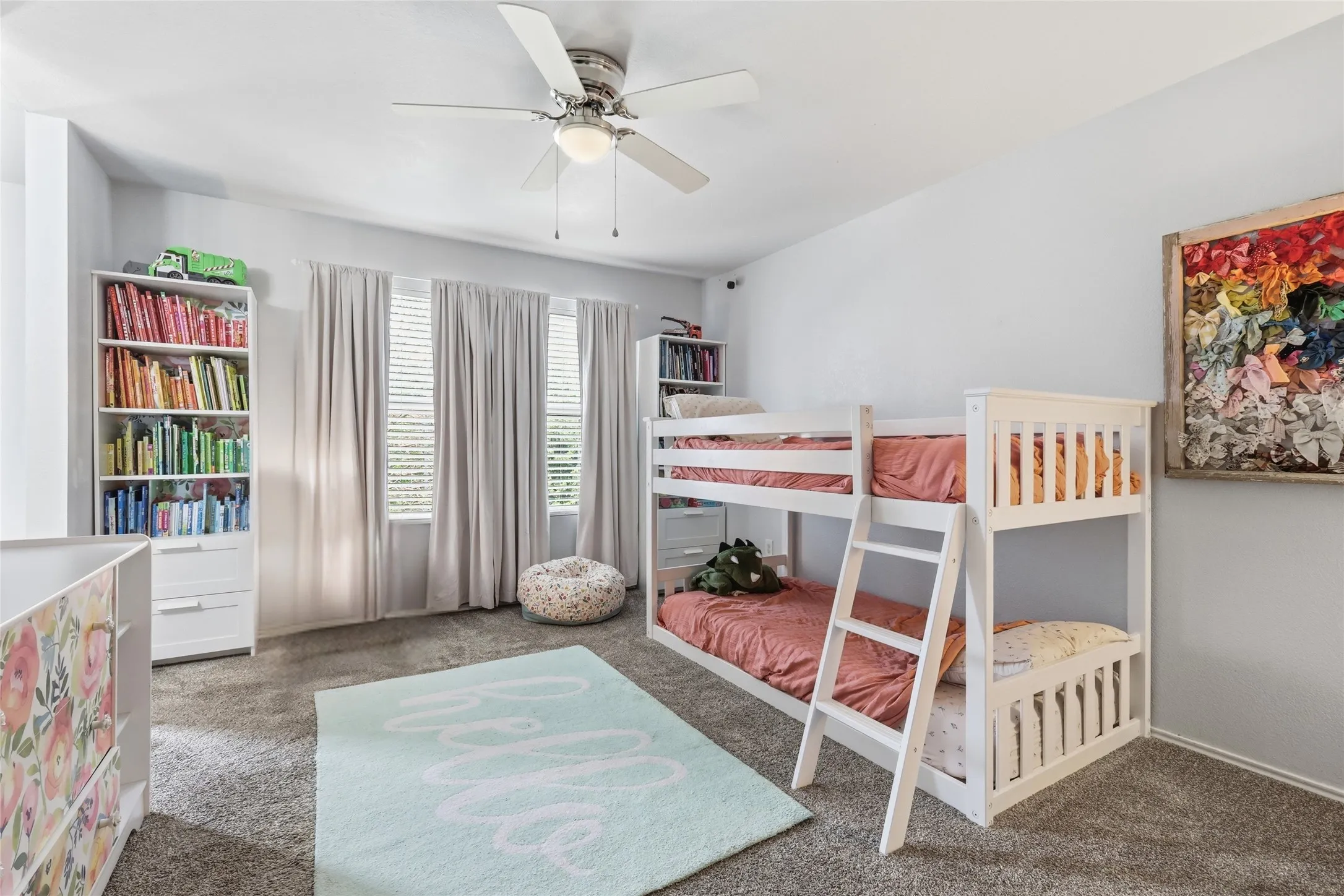 Spacious room featuring gray carpet, light gray walls, a ceiling fan with light fixture, three windows with blinds and curtains, and a built-in white bookshelf