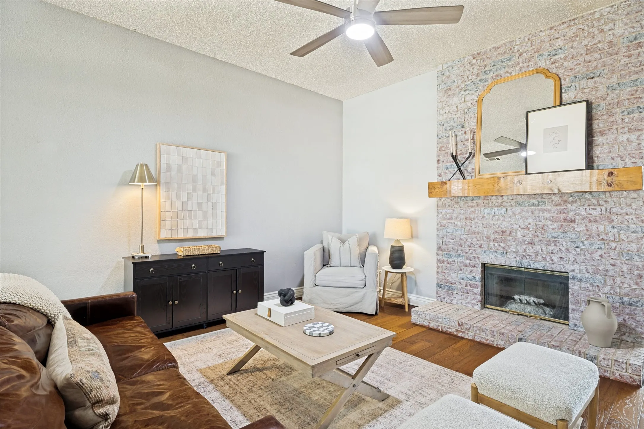 Brick-clad fireplace with a wood mantel, wood-finish flooring, and a ceiling fan with integrated lighting