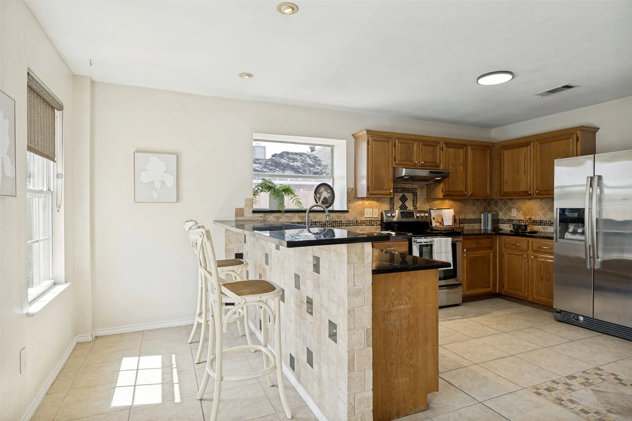 Kitchen featuring wood cabinetry, black countertops, stainless steel appliances, a tiled breakfast bar, and recessed lighting