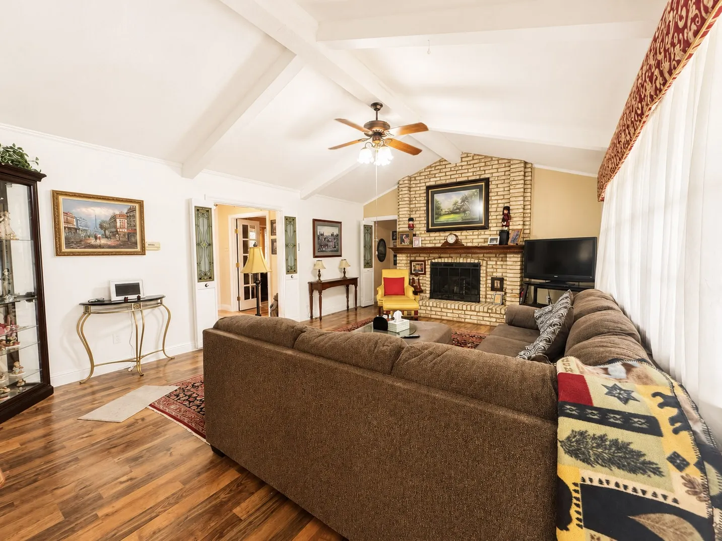 Vaulted ceiling with exposed beams, brick fireplace, wood-finish flooring, and a ceiling fan