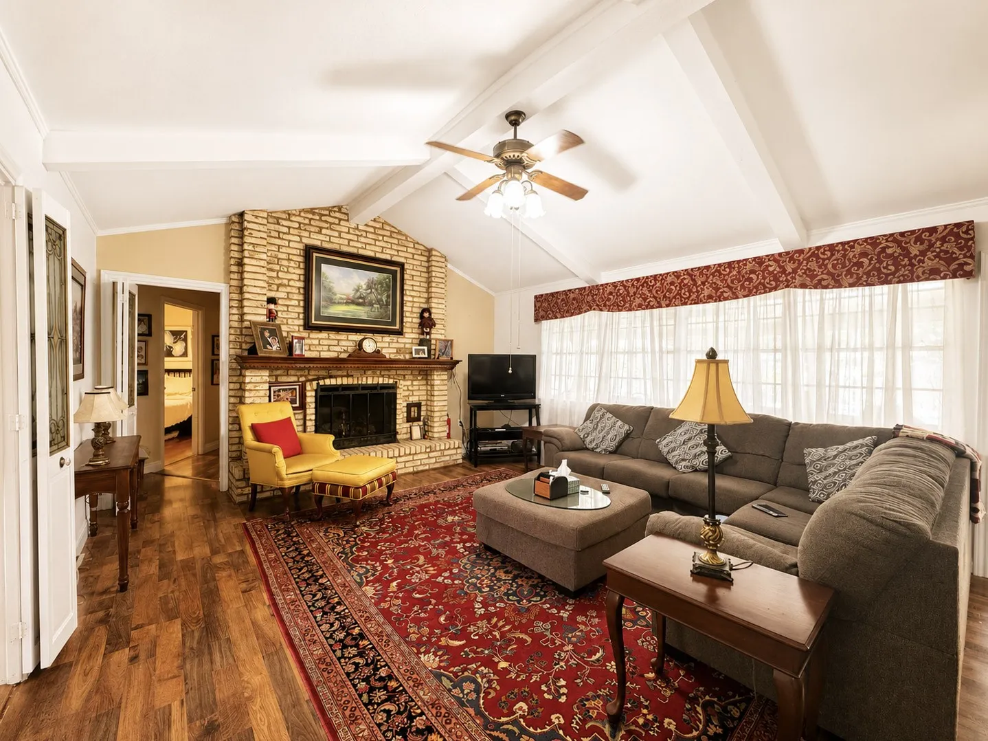 Vaulted ceiling with exposed beams, brick-clad fireplace, wood-finish flooring, and large window array