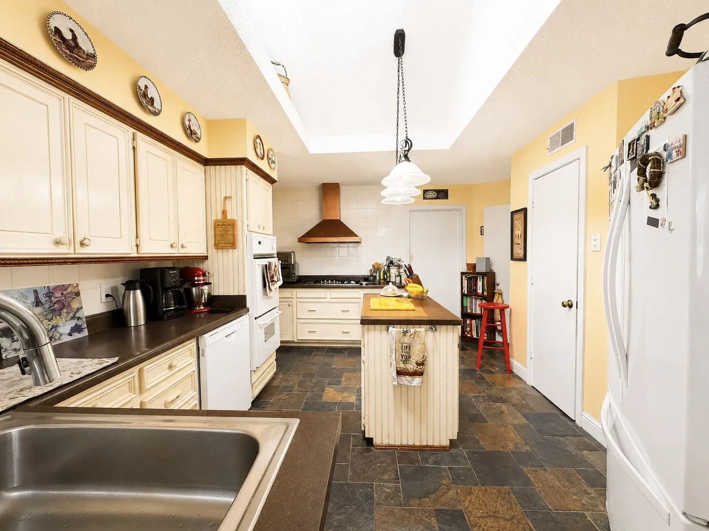 Kitchen featuring slate-style floor tile, white cabinetry with wood trim, a stainless steel sink, a built-in oven, and a copper-toned range hood