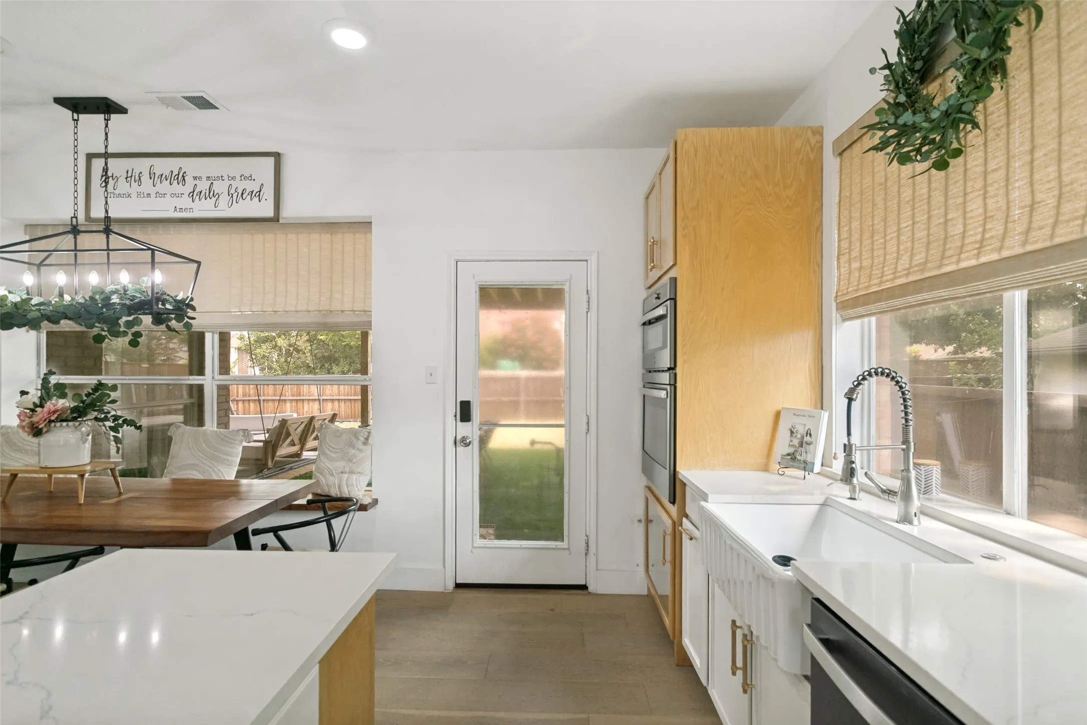 Modern kitchen featuring white cabinetry with wood-finish upper cabinets, a large white quartz island with waterfall edge, stainless steel appliances, a farmhouse-style sink with gooseneck faucet, wood-finish flooring, and abundant natural light from multiple windows and glass door opening to outdoor space