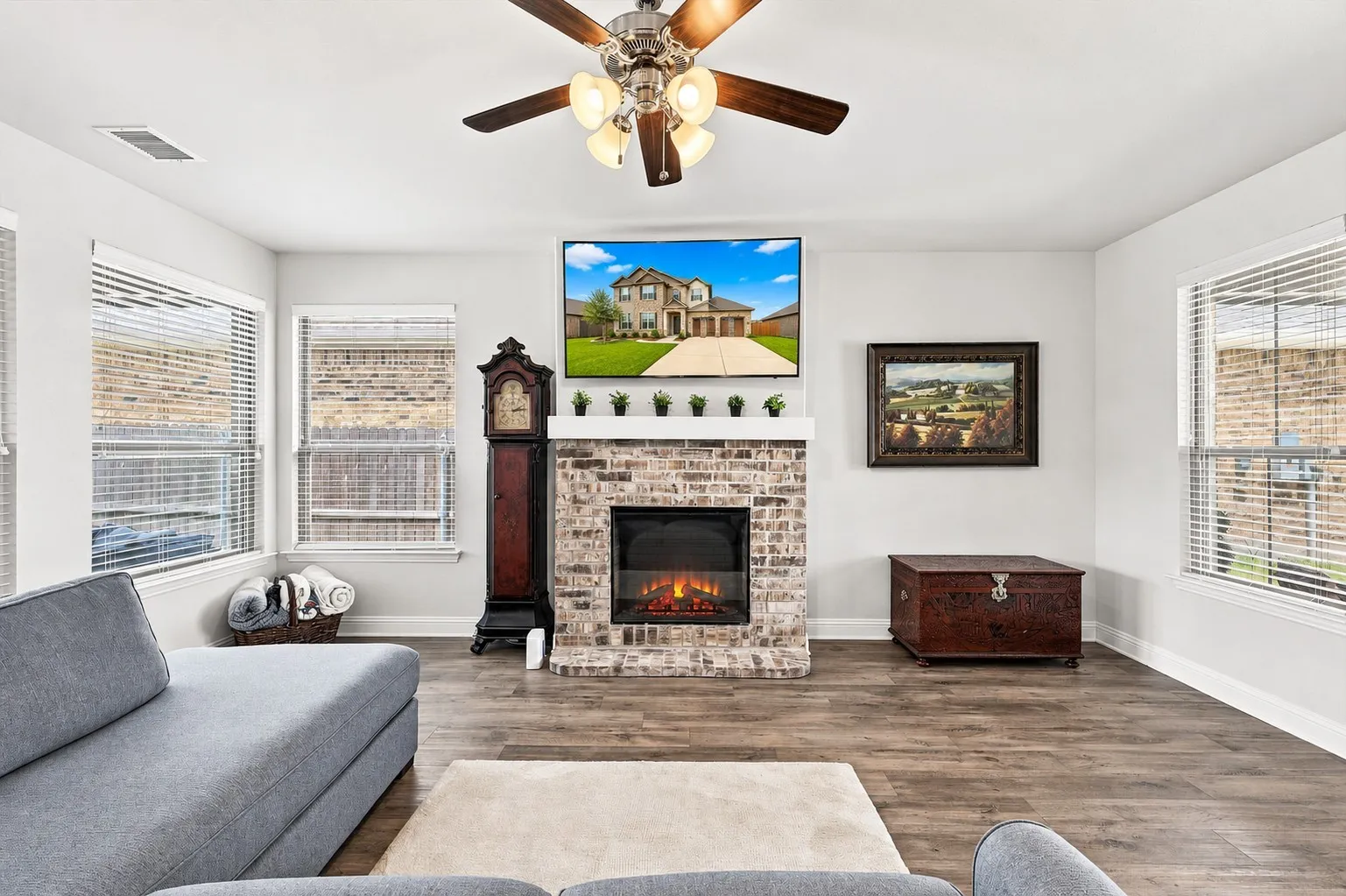 Living space featuring wood-finish flooring, a brick-surround fireplace with a mantel, multiple windows with blinds, and a ceiling fan
