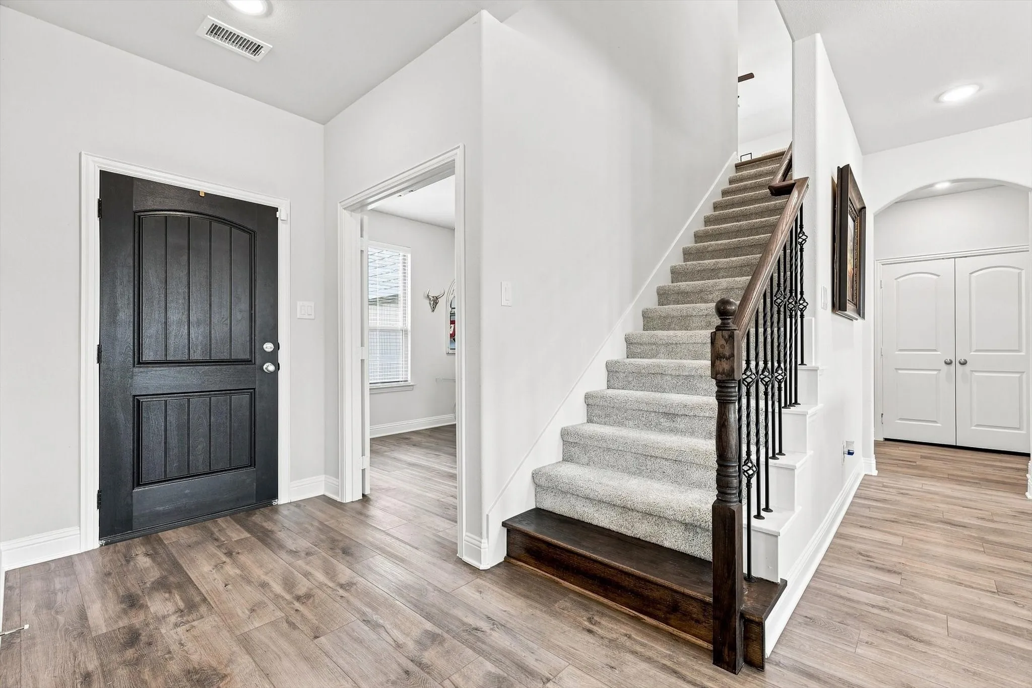 Entryway featuring wood-finish flooring, a dark wood-paneled door, and a staircase with a carpeted runner and wrought iron balusters