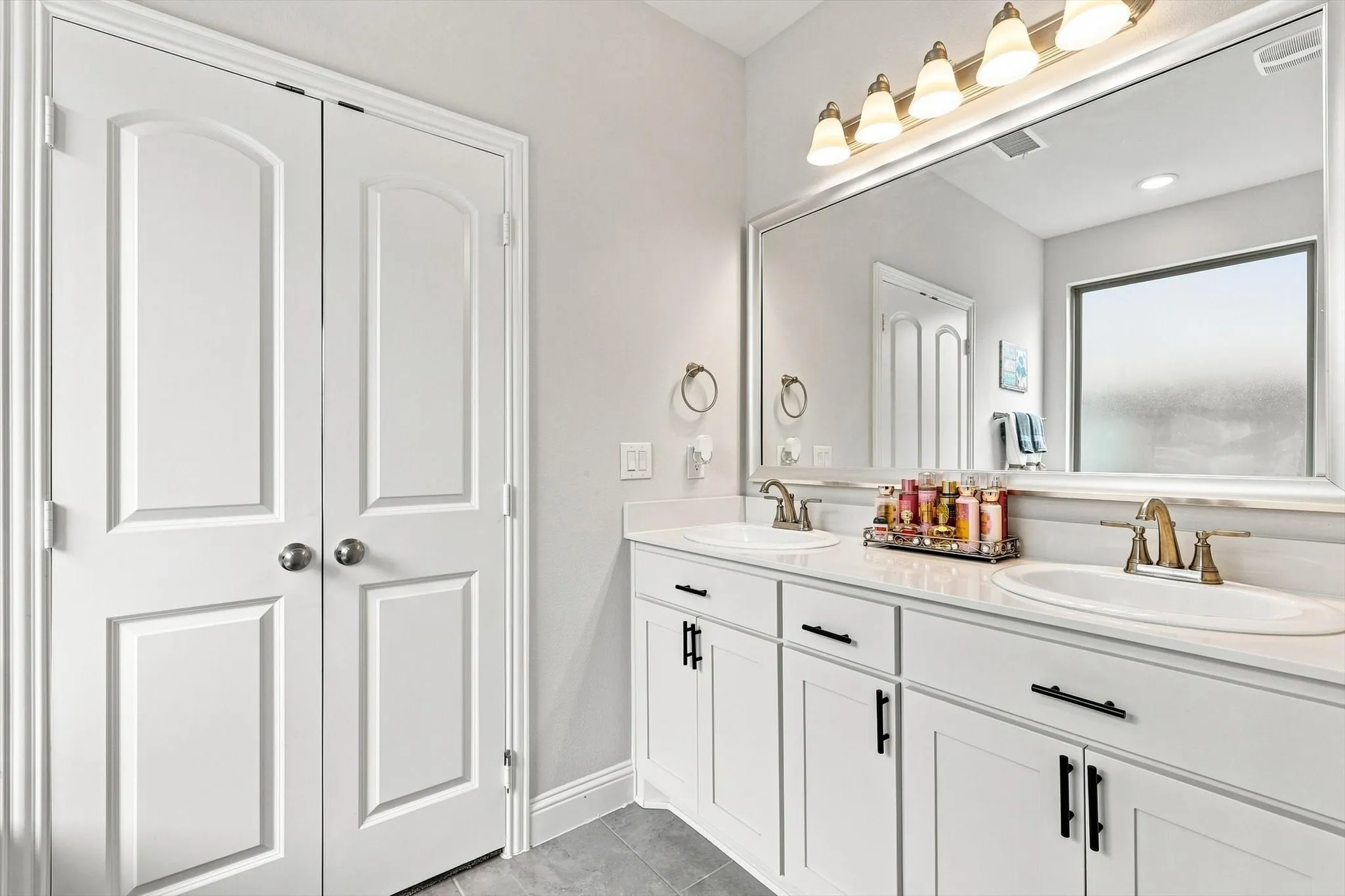 Dual white vanities with integrated sinks, brass-finish faucets, expansive framed mirror, and a multi-bulb vanity light fixture