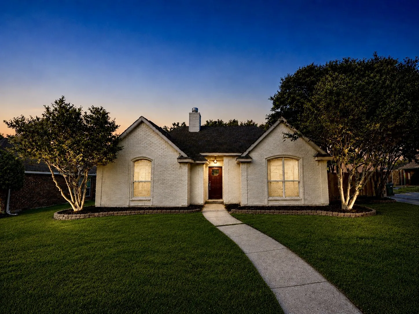 Charming painted-brick home with mature landscaping, large trees, and a warm, welcoming front entry at dusk.