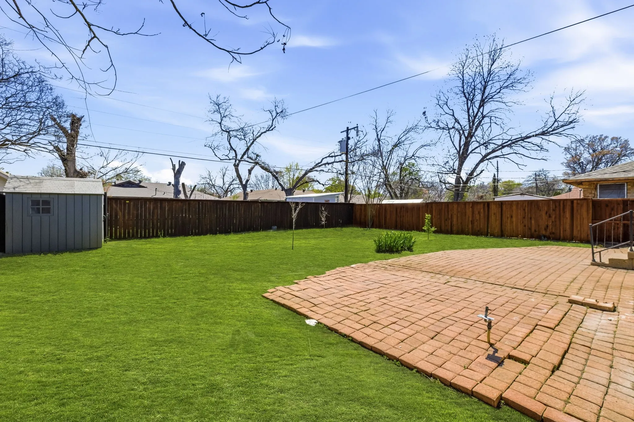 Expansive backyard featuring a manicured lawn, a red brick paver patio, a wooden privacy fence, and a storage shed
