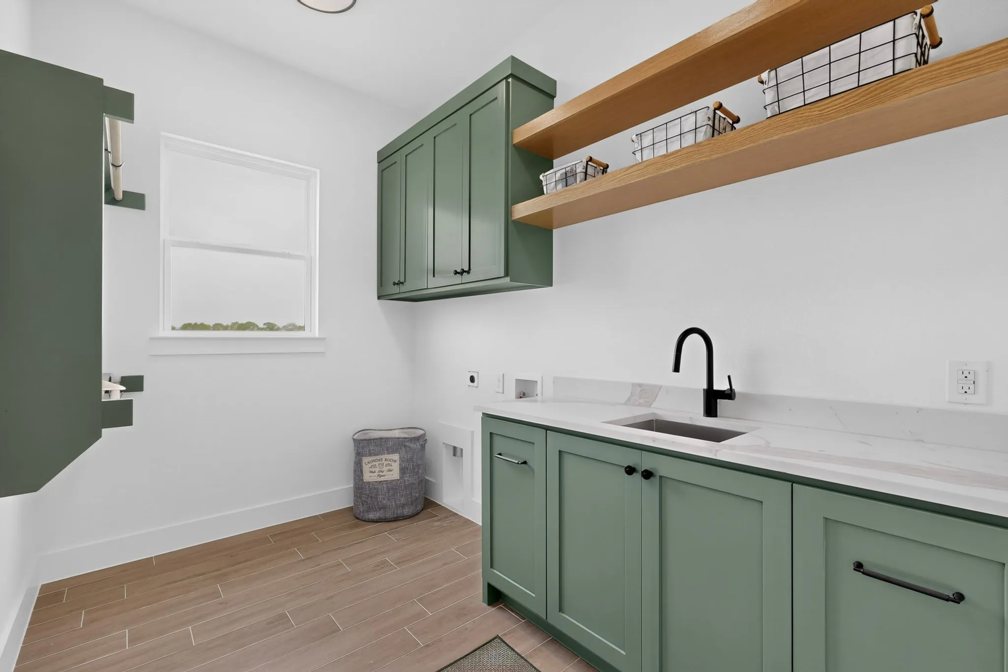 Laundry room with sage green cabinetry, a black matte faucet, light-toned countertop, and wood-finish flooring