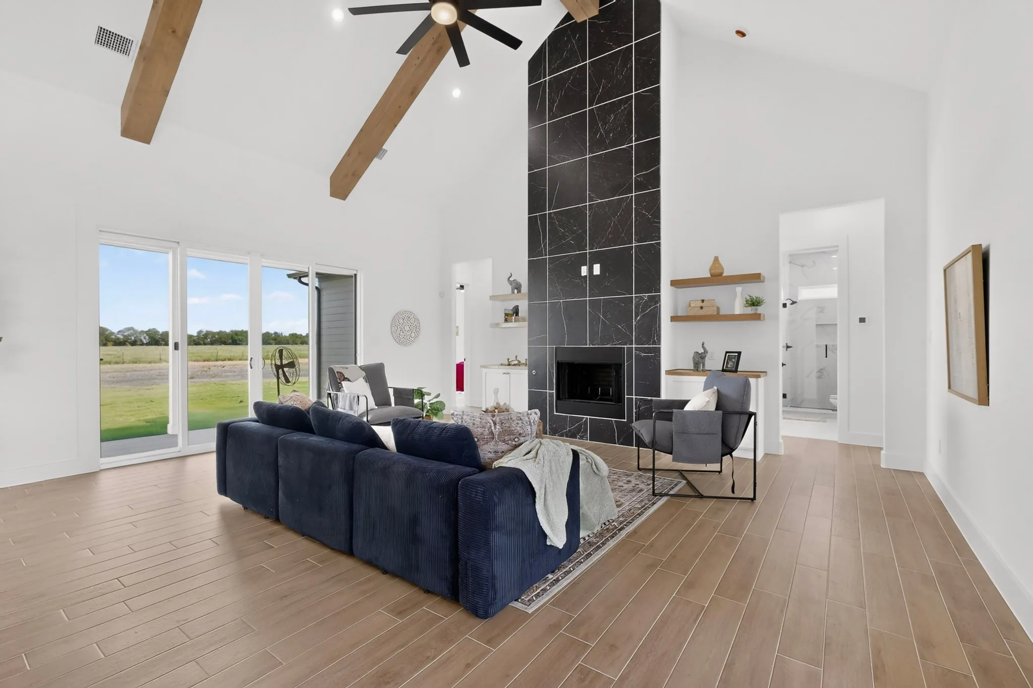 Vaulted ceiling with exposed wood beams and a ceiling fan, black tile fireplace surround with an integrated firebox, light wood-finish flooring, and white sliding glass doors