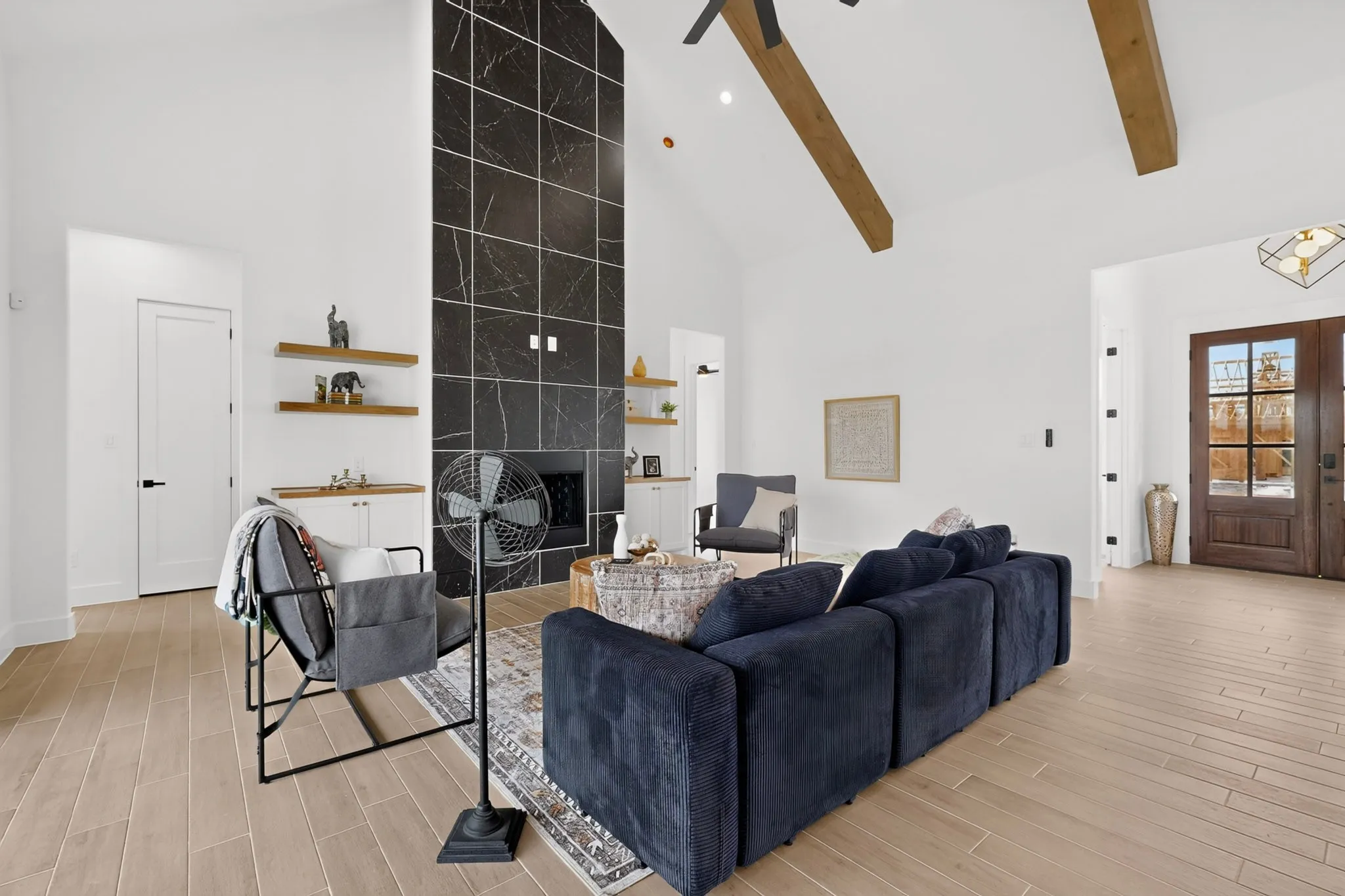 Vaulted ceiling with exposed wood beams, tall fireplace with dark-toned tile surround, light wood-finish flooring, built-in cabinetry with floating shelves, and a double-door entryway with glass inserts