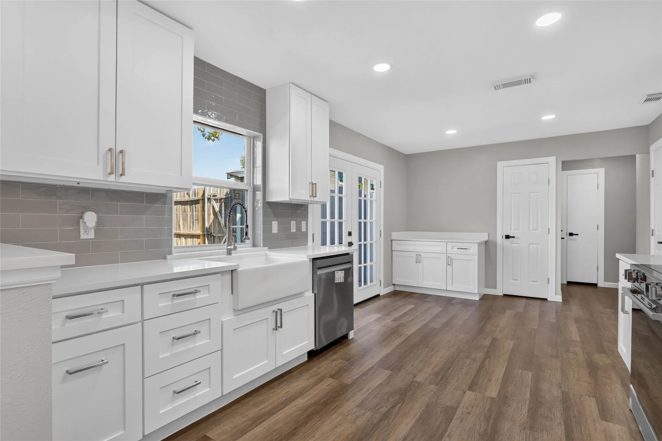 White shaker cabinetry, grey subway tile backsplash, farmhouse-style sink, stainless steel dishwasher, and wood-finish flooring