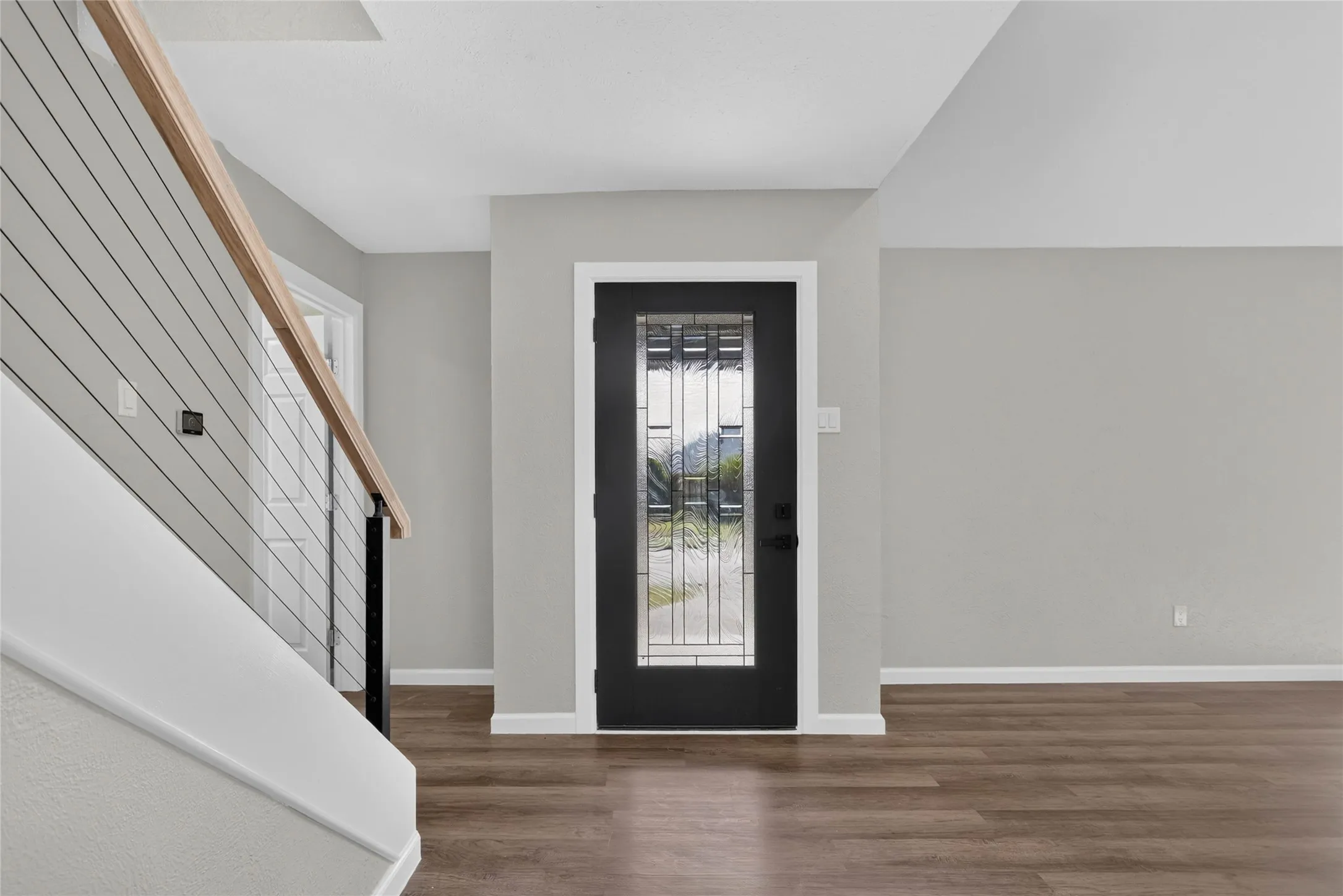 Wood-finish flooring, light gray wall tones, black entry door with decorative glass inserts, and a modern stair railing with wood handrail