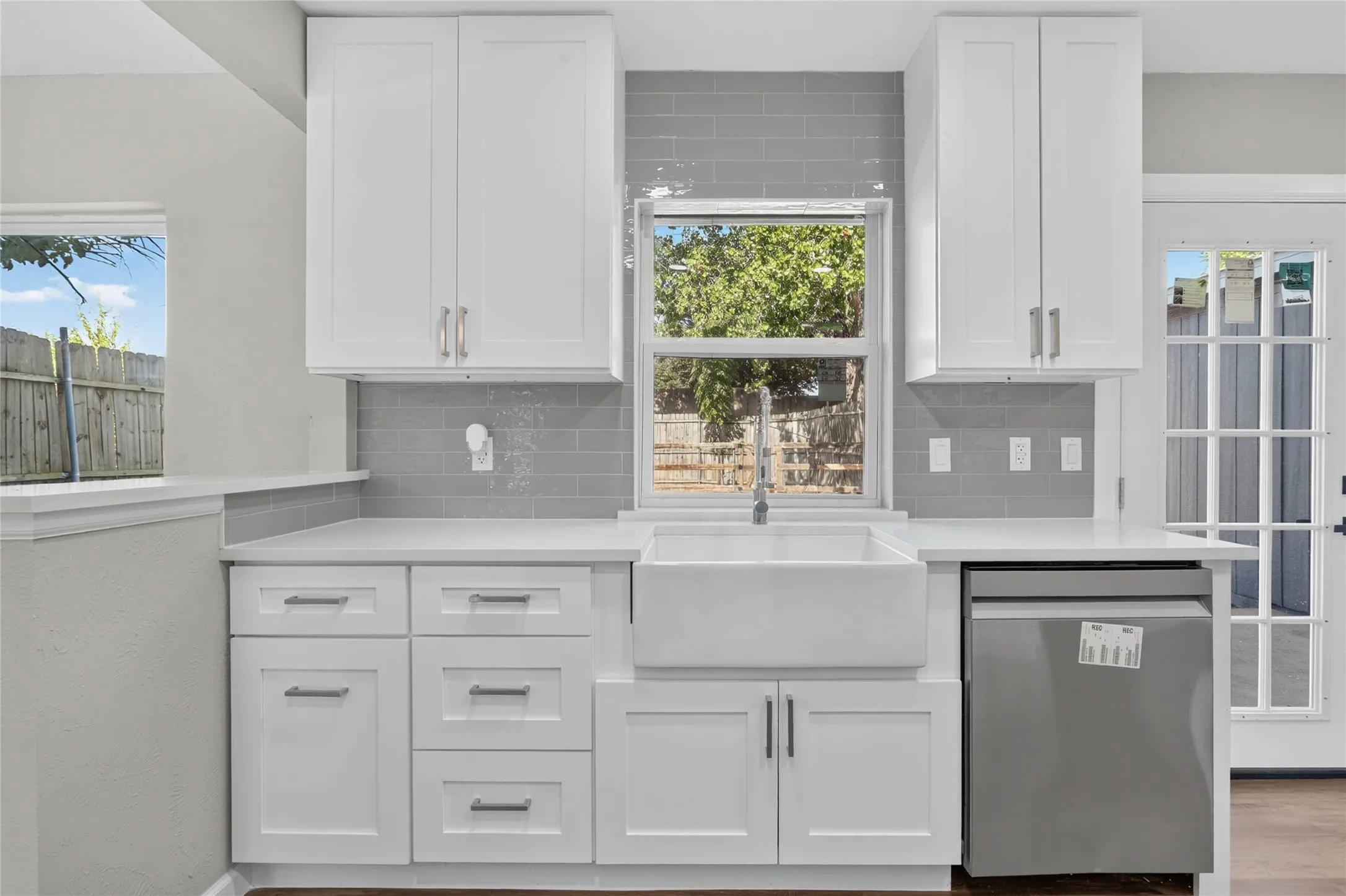 White shaker cabinetry with modern hardware, white apron-front sink, light-toned countertops, gray subway tile backsplash, and stainless steel dishwasher