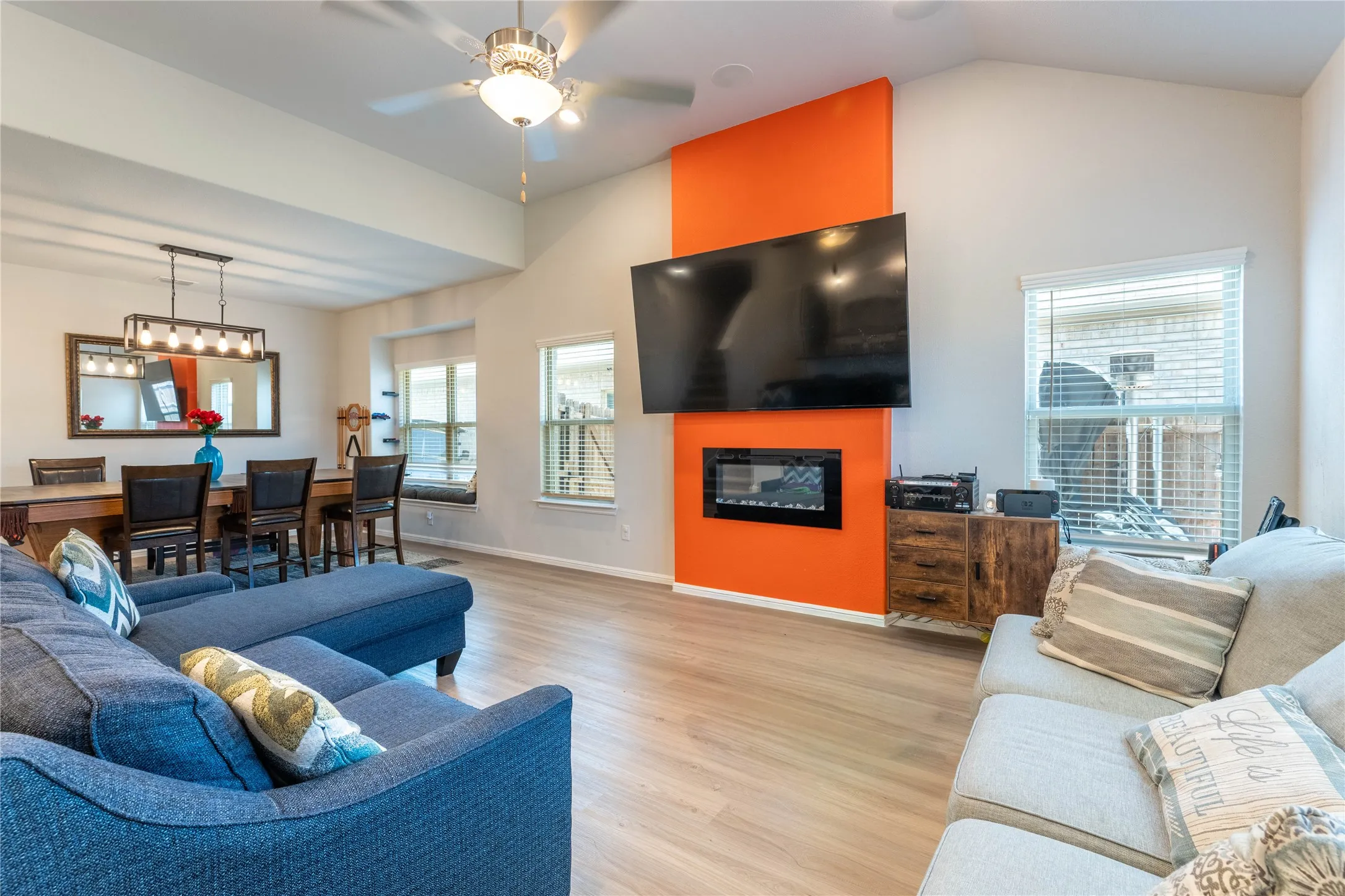 Vaulted ceiling with ceiling fan, wood-finish flooring, accent wall with built-in electric fireplace, recessed lighting, and dining area with linear pendant light
