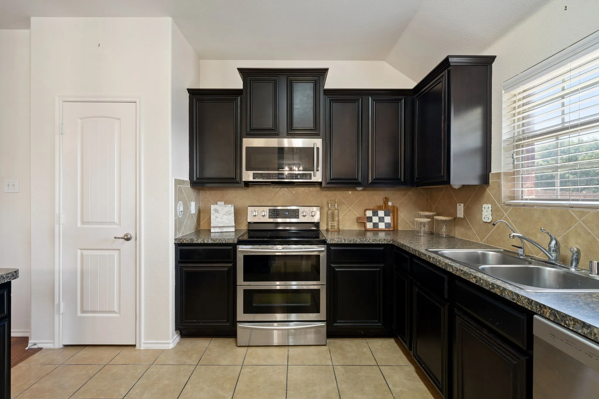 Kitchen featuring dark wood-finish cabinetry, stainless steel appliances, speckled countertops, a tile backsplash, and tile flooring