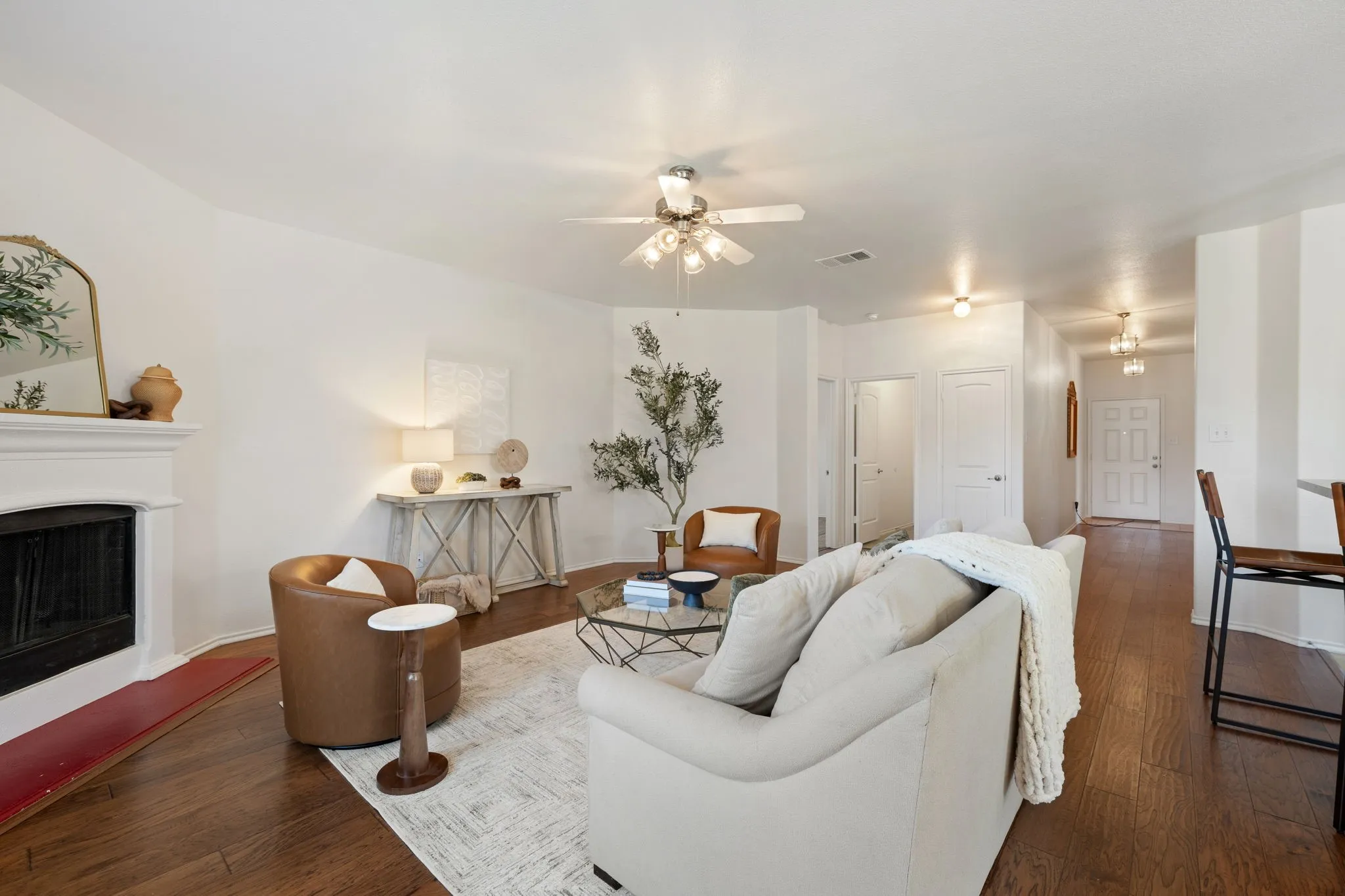 Hardwood-finish flooring, white painted walls, fireplace with white mantel, ceiling fan with integrated lighting, and multiple light fixtures