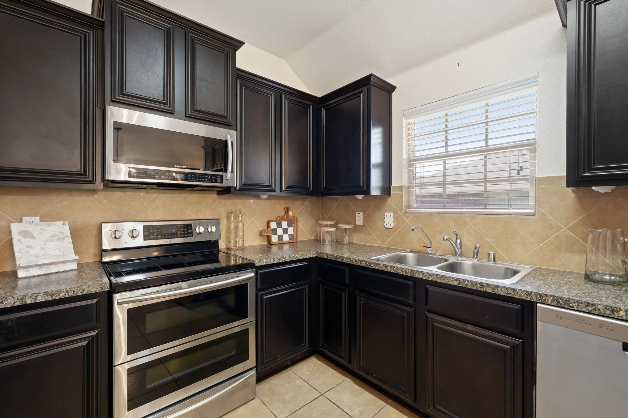 Dark wood-finish cabinetry, stainless steel appliances, stone-finish countertops, tile backsplash, and a double basin sink