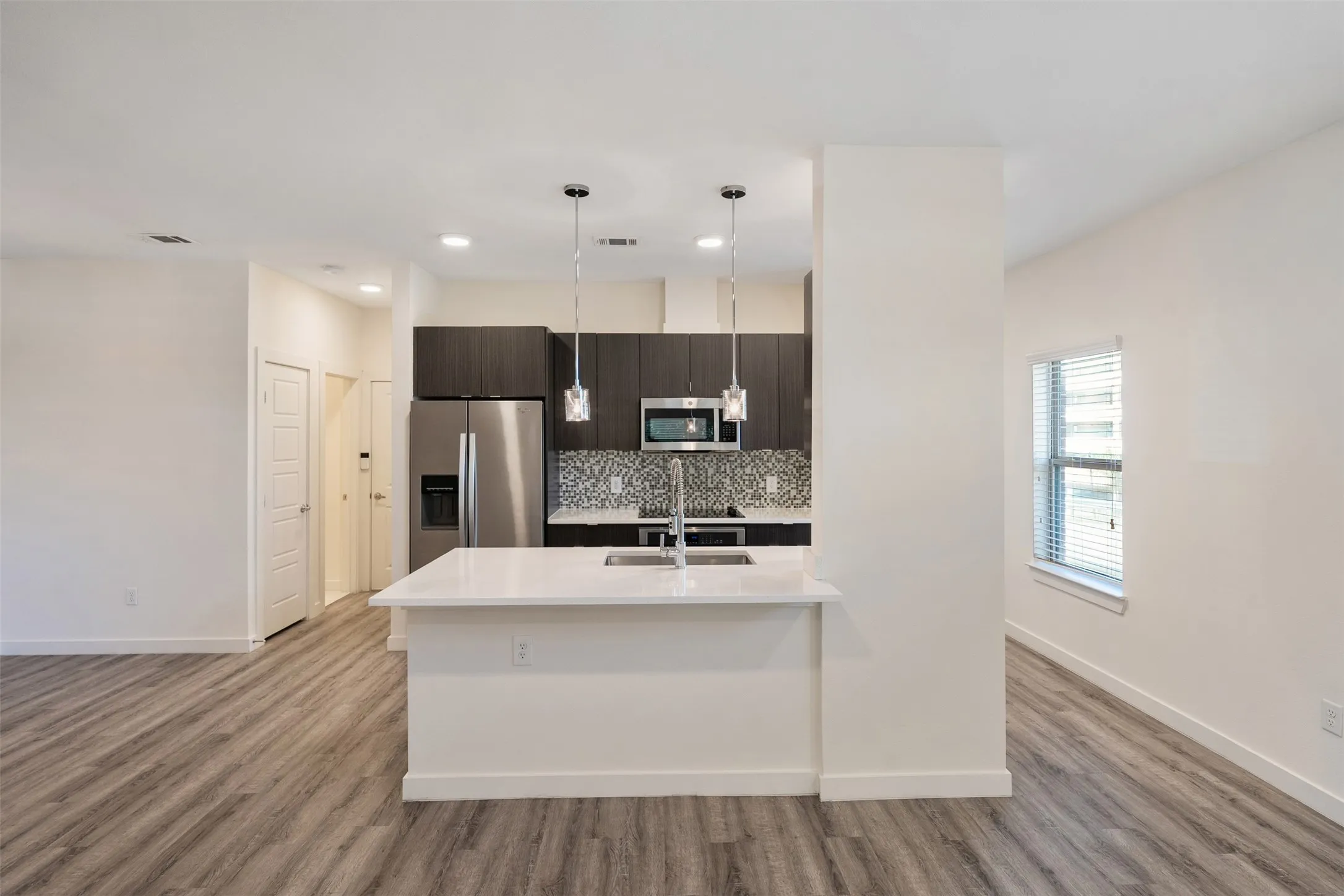 Kitchen island with a white countertop and integrated sink, stainless steel appliances, dark wood-finish cabinetry, patterned tile backsplash, and wood-finish flooring