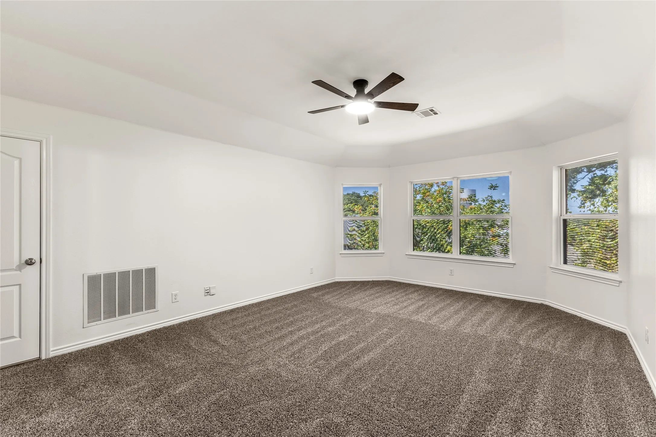Spacious room featuring neutral carpeting, white walls, multiple windows, a ceiling fan, and a paneled door