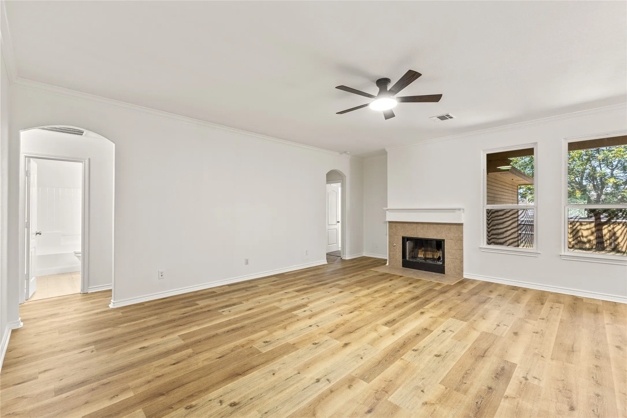 Spacious room featuring wood-finish flooring, a ceiling fan with integrated lighting, a fireplace with a stone surround, and multiple windows