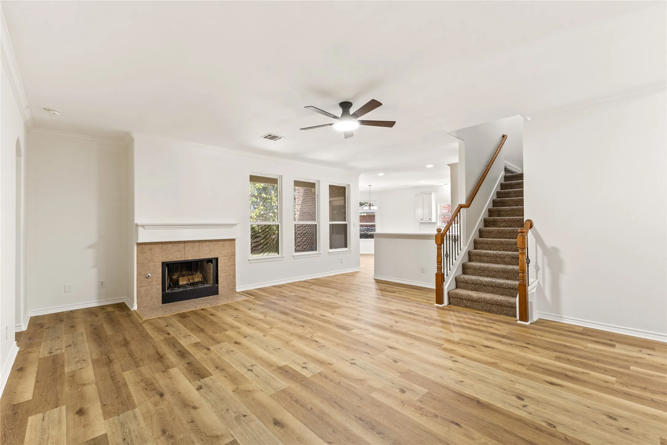 Spacious living area featuring wood-finish flooring, a tiled fireplace, multiple windows, a ceiling fan, and a carpeted staircase with wood banister
