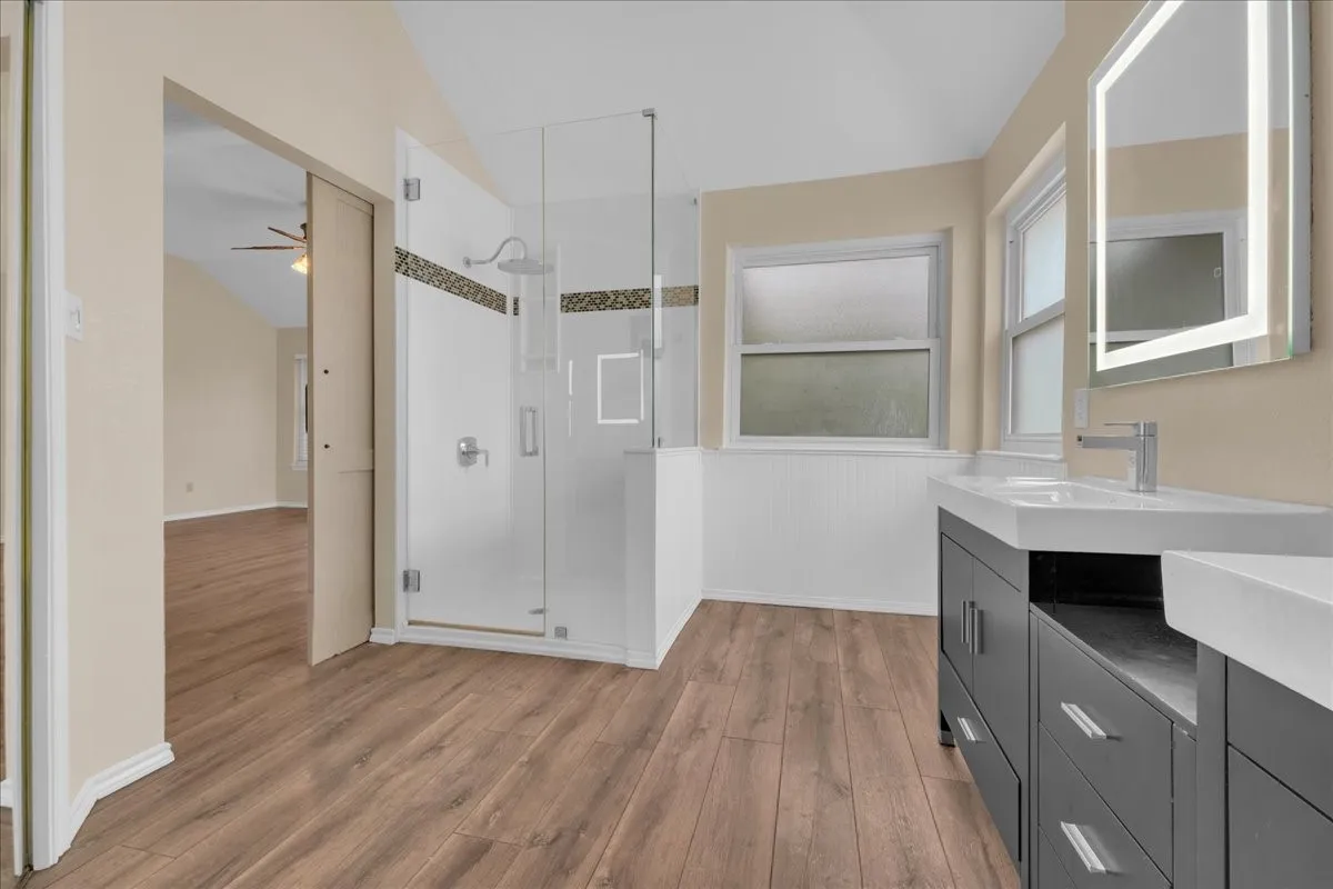 Wood-finish flooring throughout, glass-enclosed shower with tile accent, dual vanity with undermount sinks, illuminated mirrors, and panel wainscoting