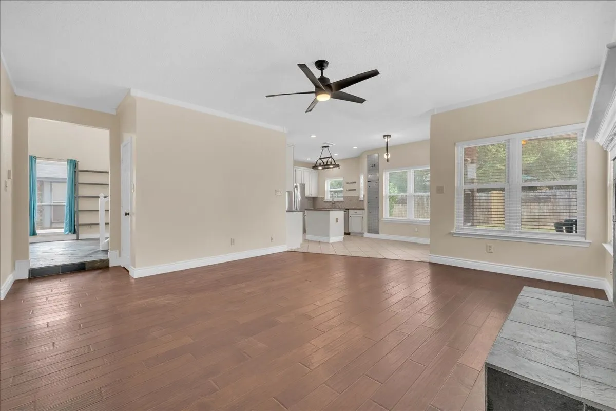 Wood-finish flooring throughout, ceiling fan with integrated lighting, multiple windows providing natural light, white kitchen cabinetry, and a tiled fireplace surround